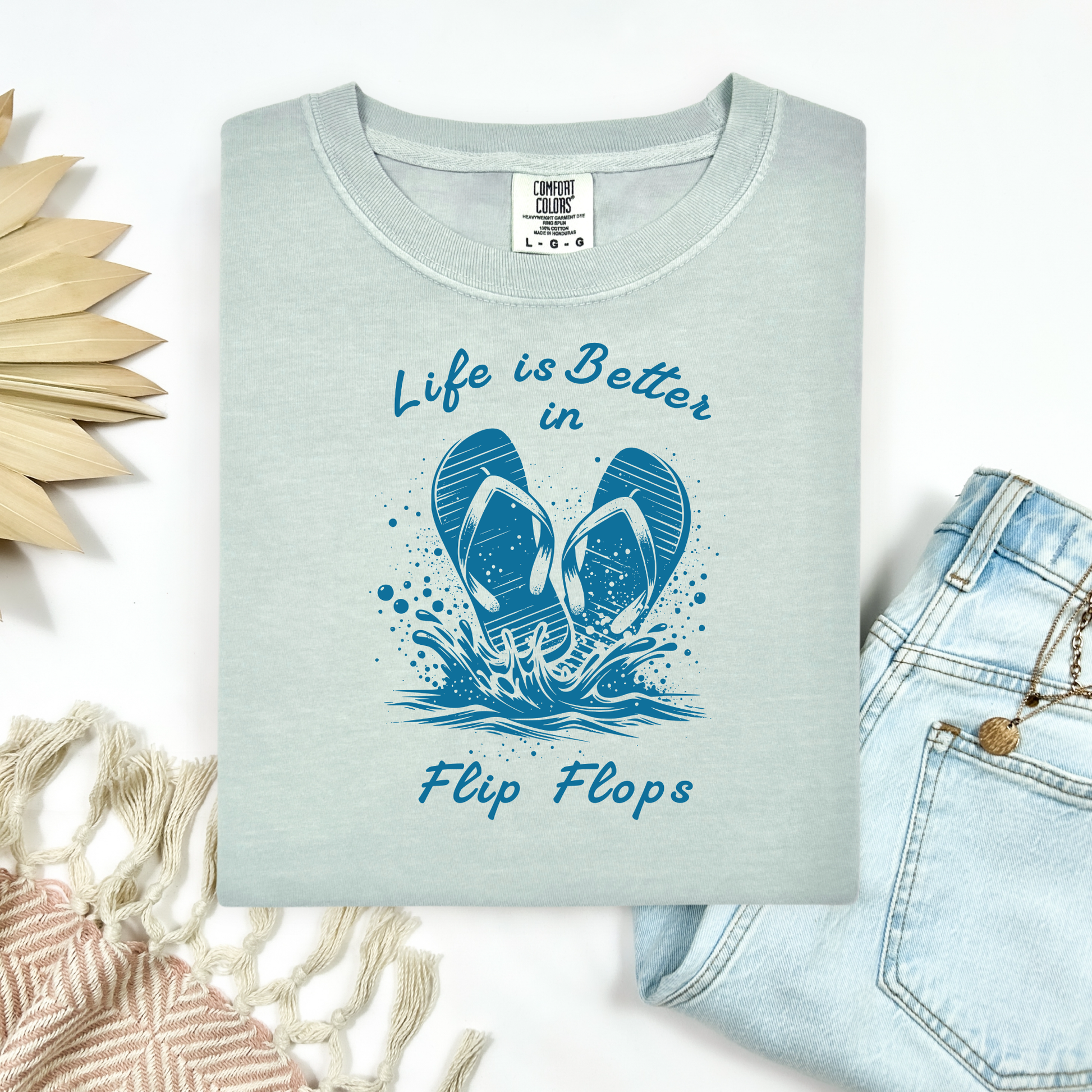 Comfort Colors 1717 "Life is Better in Flip Flops" t shirt with a pair of flip flops splashing in water one color graphic. Picture on a flay lay mockup with pair of blue jeans and blanket and a wicker sun shaped accessory.