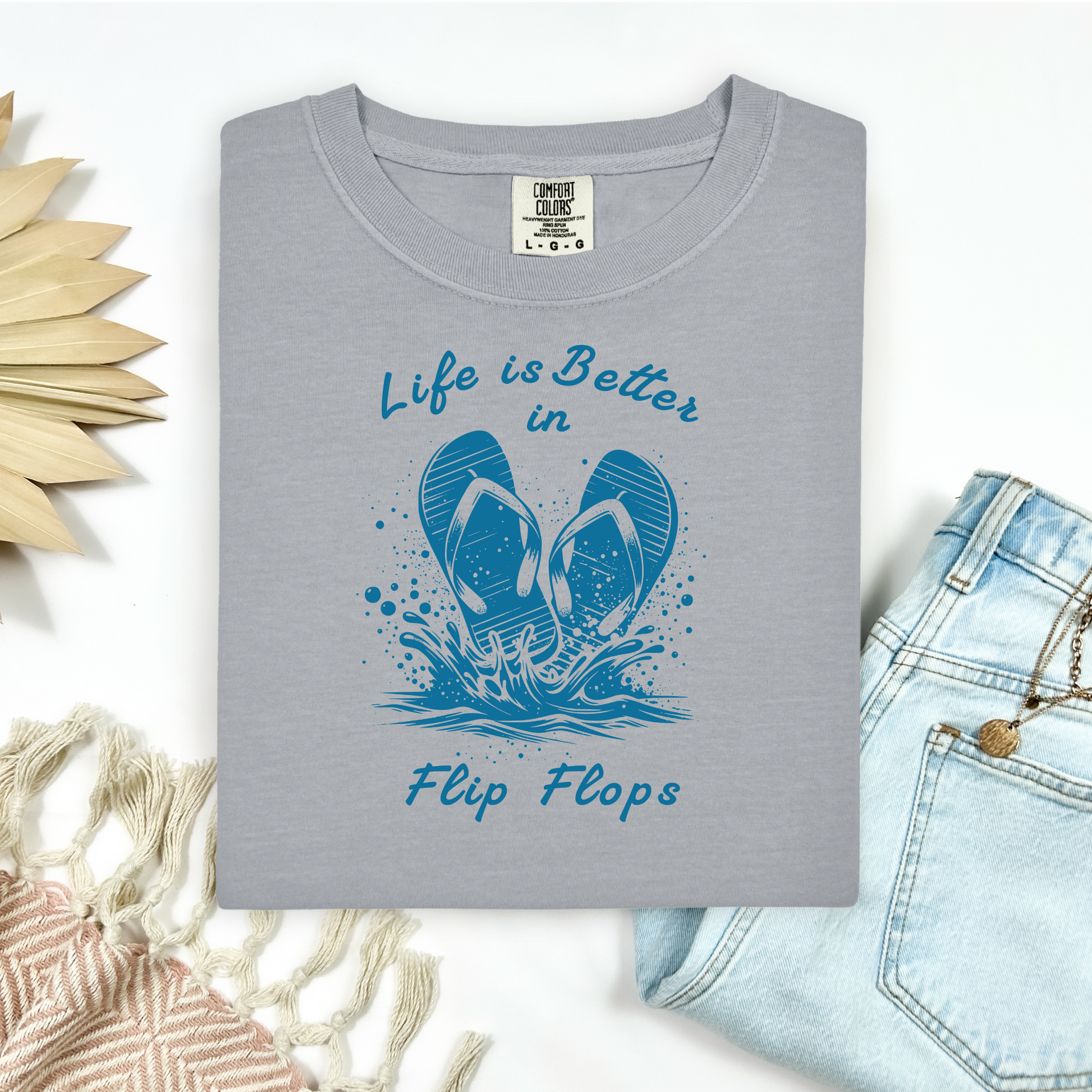Comfort Colors 1717 "Life is Better in Flip Flops" t shirt with a pair of flip flops splashing in water one color graphic. Picture on a flay lay mockup with pair of blue jeans and blanket and a wicker sun shaped accessory.