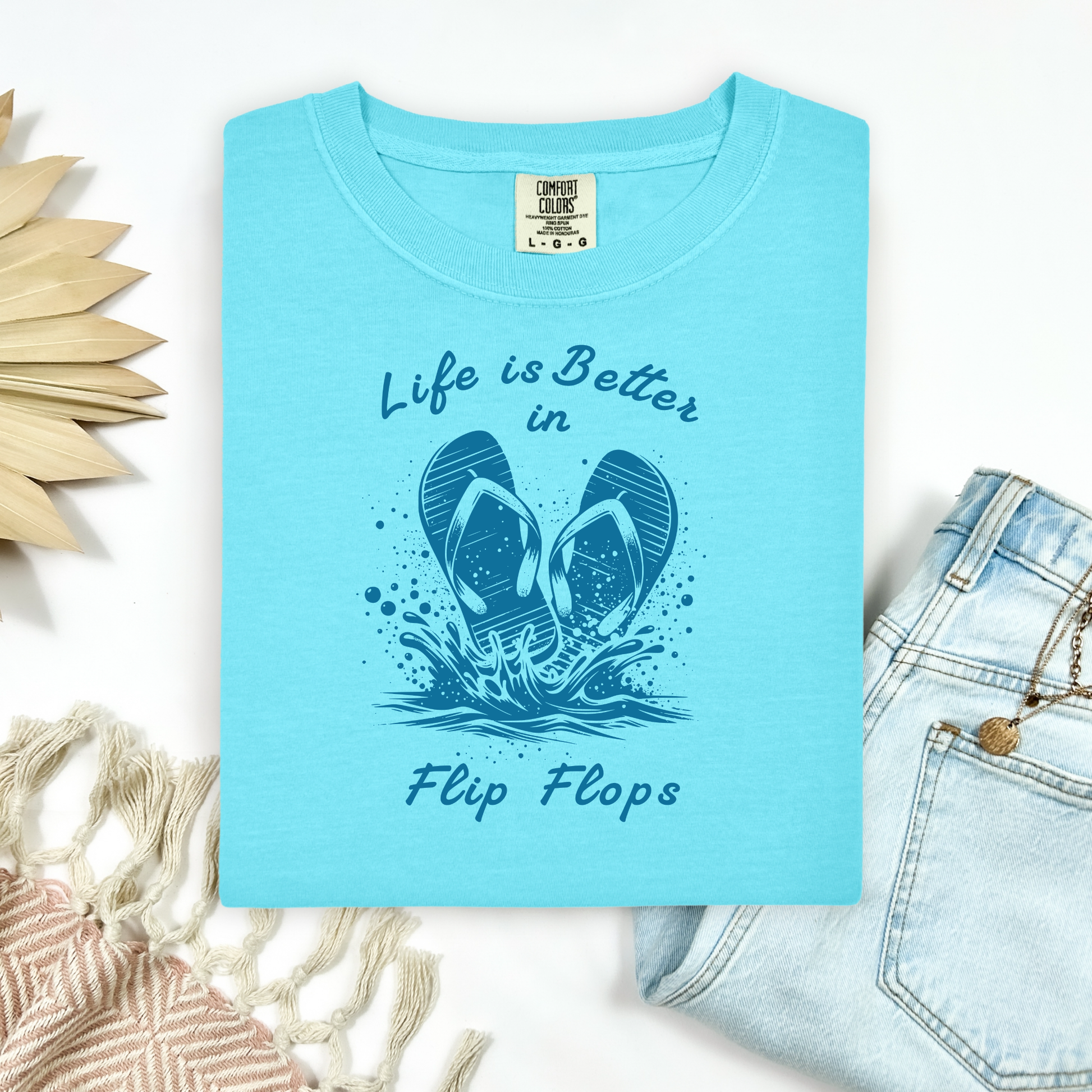 Comfort Colors 1717 "Life is Better in Flip Flops" t shirt with a pair of flip flops splashing in water one color graphic. Picture on a flay lay mockup with pair of blue jeans and blanket and a wicker sun shaped accessory.