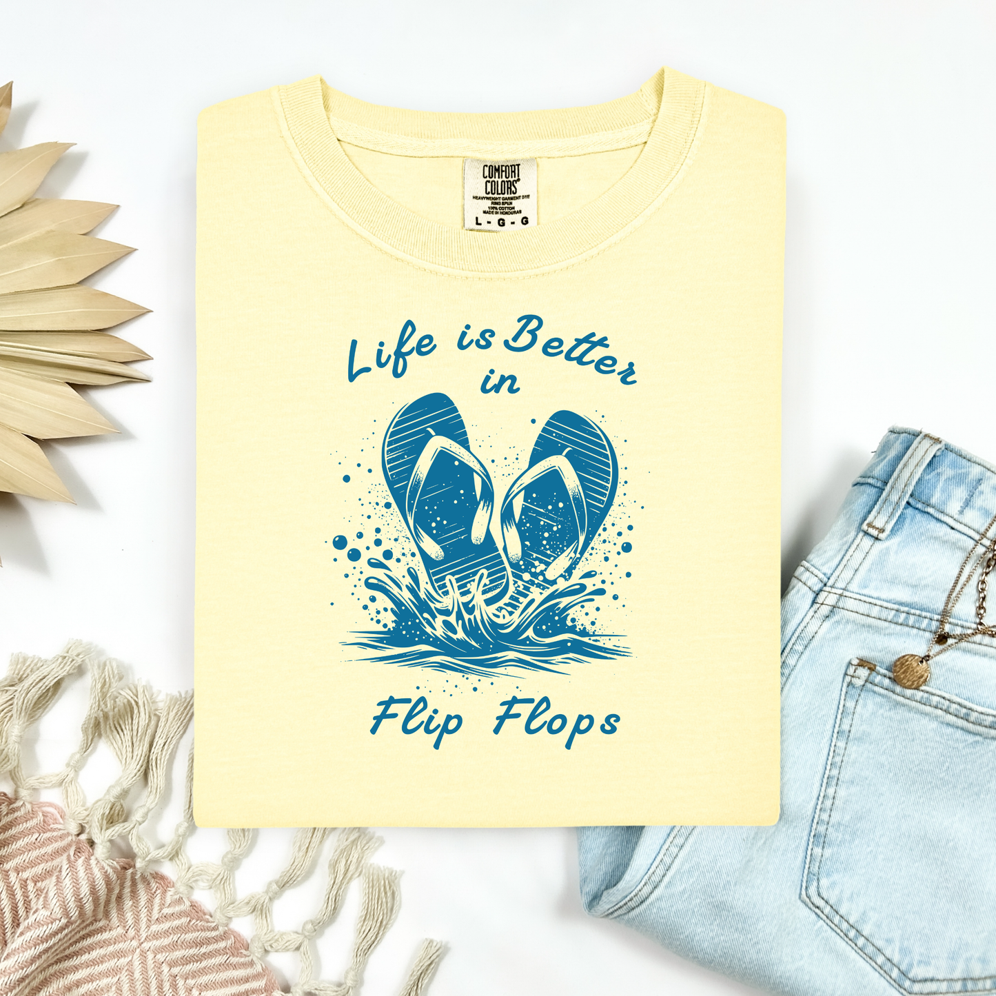Comfort Colors 1717 "Life is Better in Flip Flops" t shirt with a pair of flip flops splashing in water one color graphic. Picture on a flay lay mockup with pair of blue jeans and blanket and a wicker sun shaped accessory.