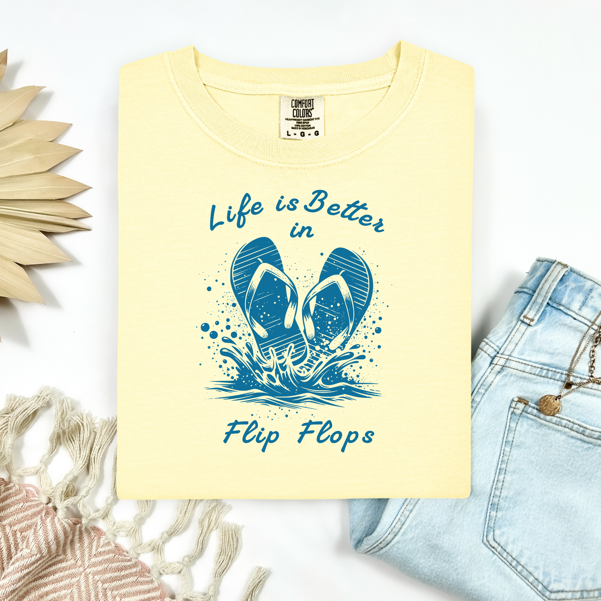 Comfort Colors 1717 "Life is Better in Flip Flops" t shirt with a pair of flip flops splashing in water one color graphic. Picture on a flay lay mockup with pair of blue jeans and blanket and a wicker sun shaped accessory.