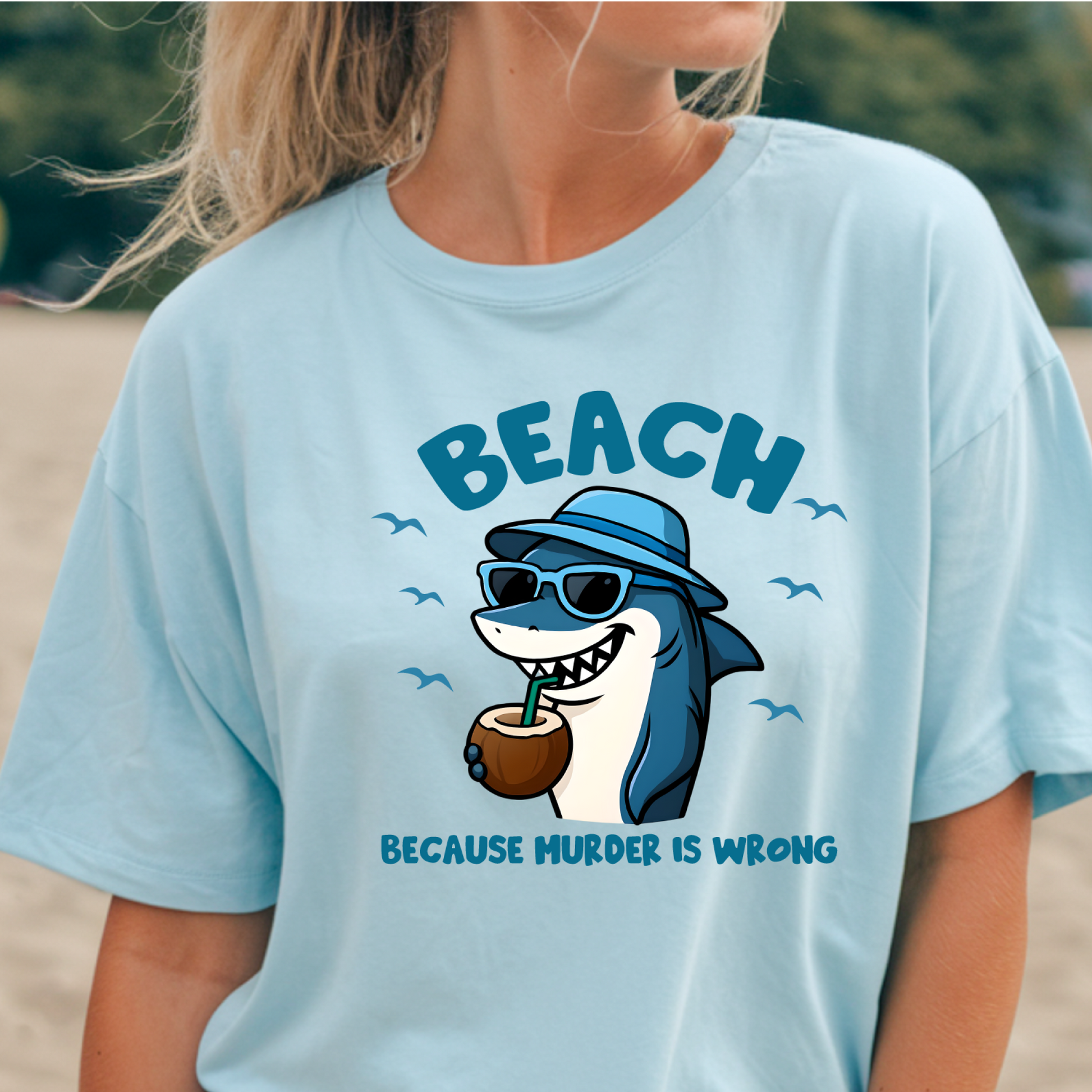 Light blue t-shirt with a shark graphic and text on a beach.