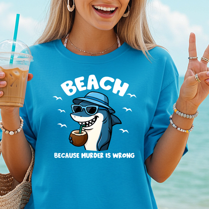 Person wearing a blue t-shirt with a shark graphic and text, holding a drink by the beach.