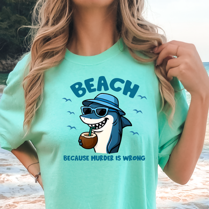 Person wearing a mint green t-shirt with a shark graphic and text at a beach.