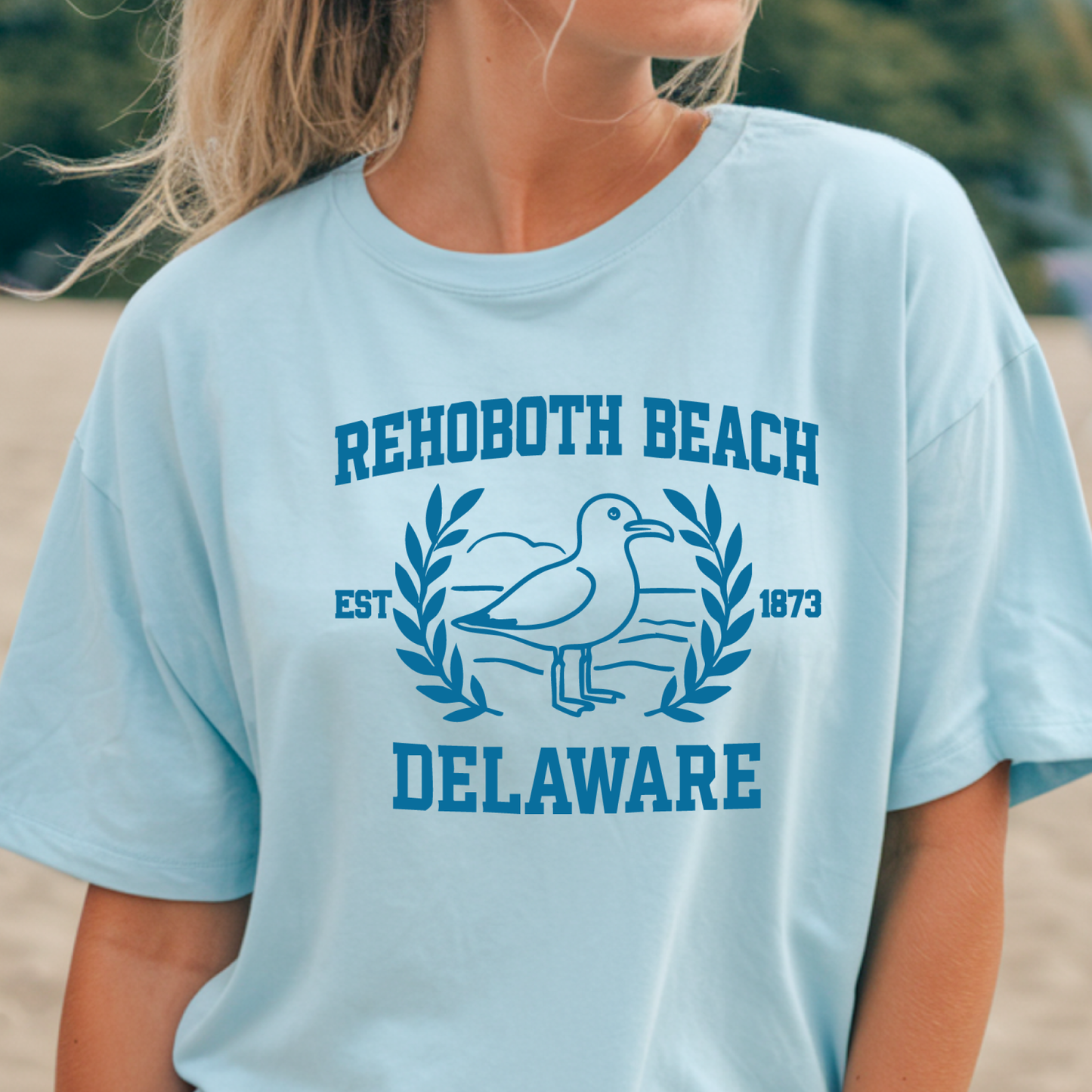 Light blue t-shirt with Rehoboth Beach Delaware design on a blurred beach background