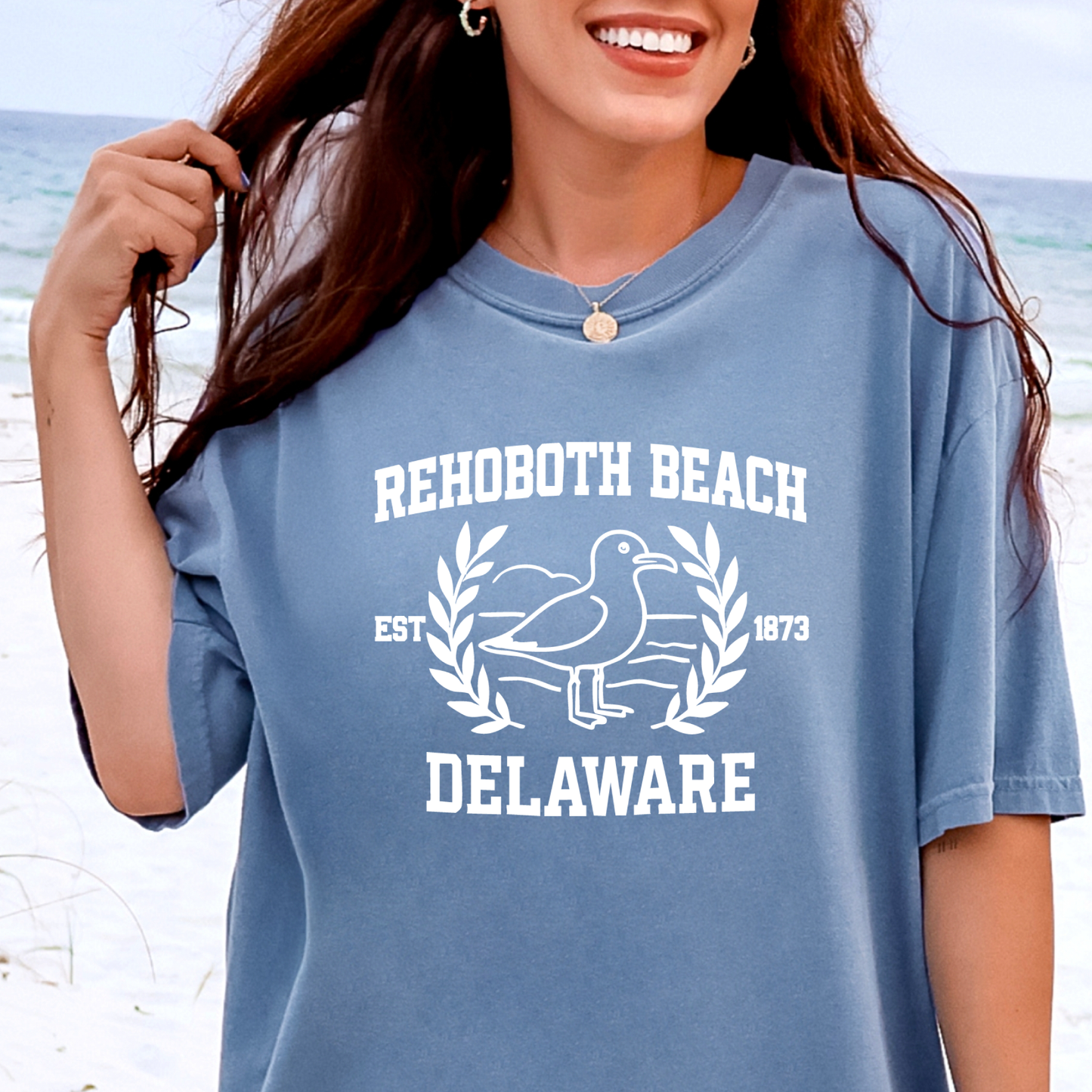 Person wearing a blue t-shirt with Rehoboth Beach, Delaware text and graphics on a beach background