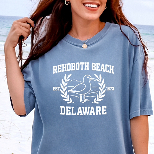 Person wearing a blue t-shirt with Rehoboth Beach, Delaware text and graphics on a beach background
