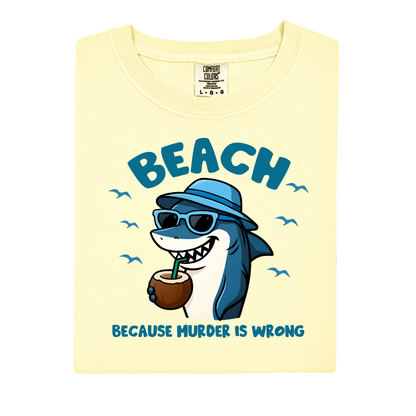 Shirt with a cartoon shark wearing sunglasses and a hat, holding a coconut, and text 'BEACH BECAUSE MURDER IS WRONG' on a light yellow background.