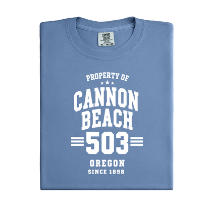 Folded blue t-shirt with 'Property of Cannon Beach 503 Oregon Since 1898' text on a white background
