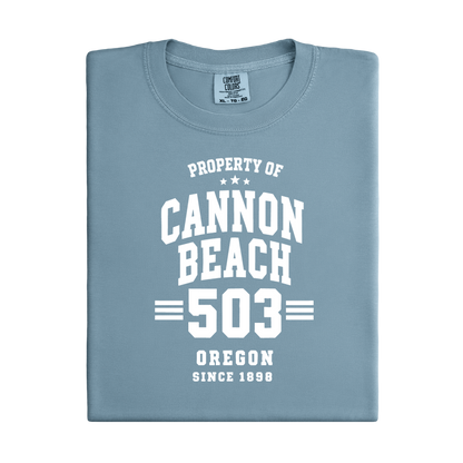 Folded blue t-shirt with white text 'Property of Cannon Beach 503 Oregon Since 1899' on a white background