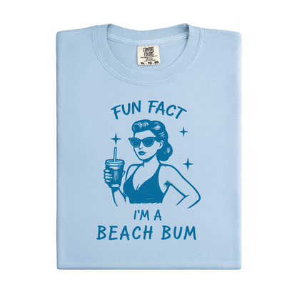 Light blue t-shirt with a graphic of a woman in a bikini and sunglasses, holding a drink, with text 'Fun Fact I'm a Beach Bum' on a white background.
