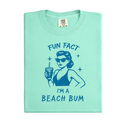 Light green t-shirt with a graphic of a woman in a bikini and sunglasses, holding a drink, with text 'Fun Fact I'm a Beach Bum' on a white background.