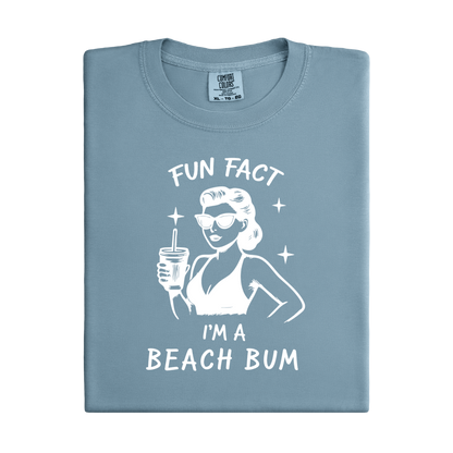 Folded blue t-shirt with a graphic of a woman in a bikini holding a drink and text 'Fun Fact I'm a Beach Bum' on a white background.