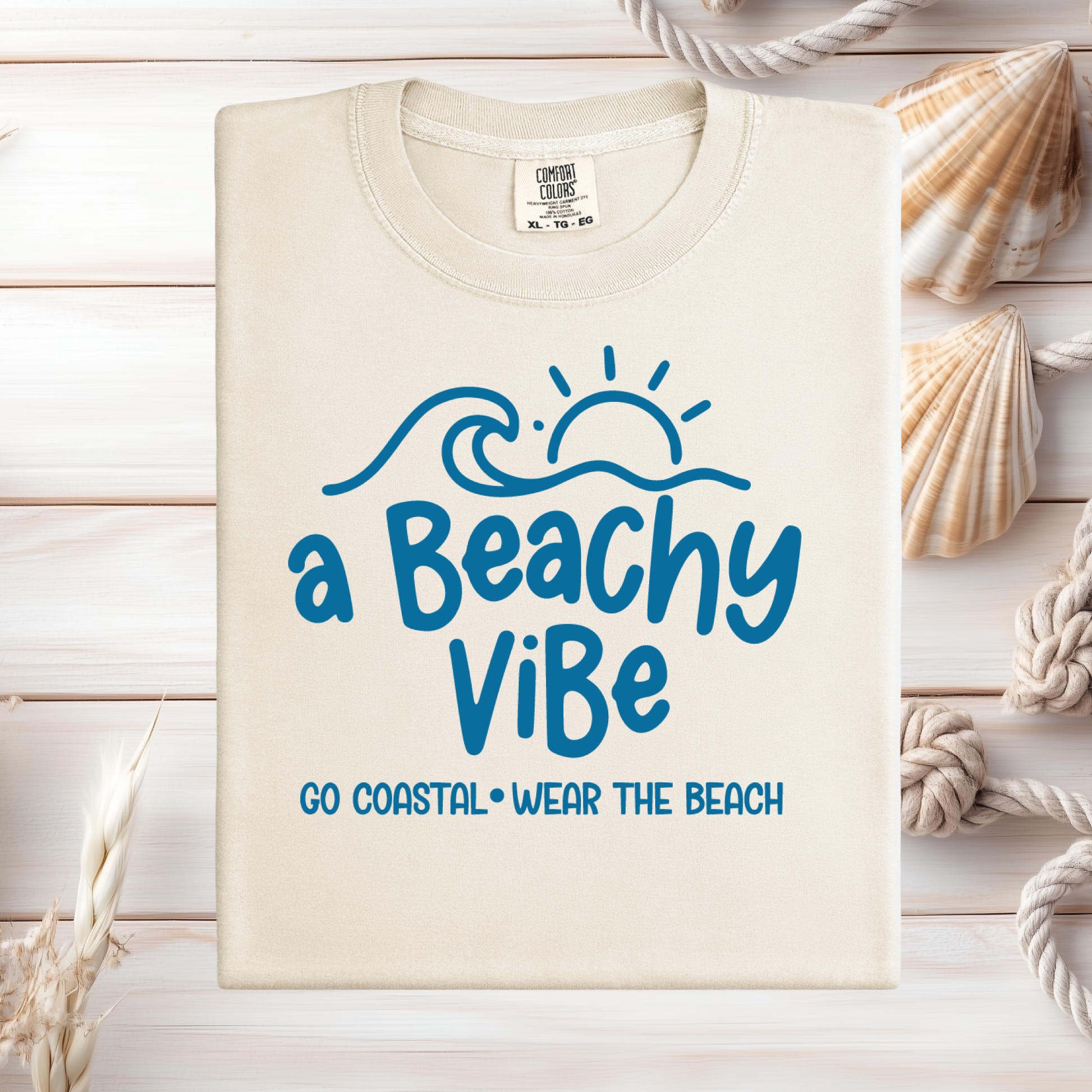 Beige t-shirt with 'a Beachy Vibe' text on a wooden surface with seashells and rope.