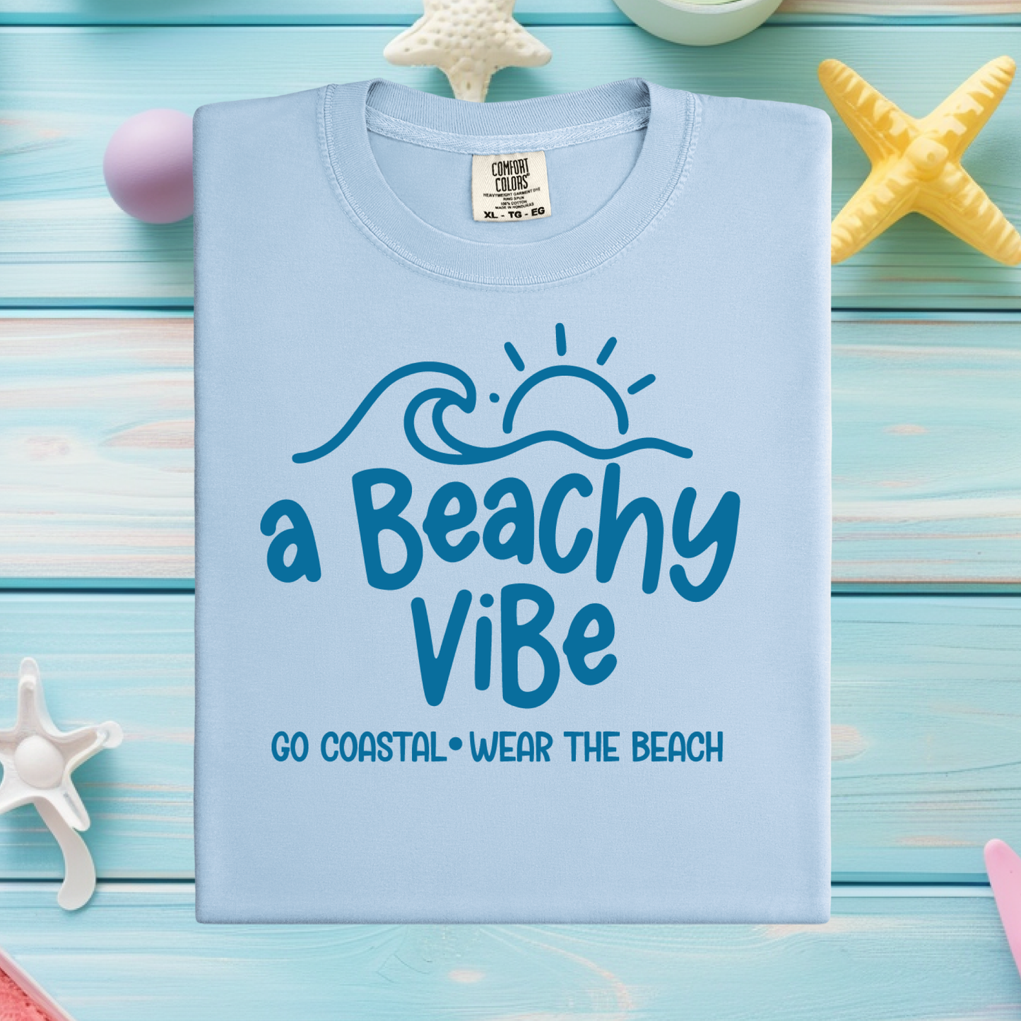 Light blue t-shirt with 'a Beachy ViBe' text on a wooden surface with starfish decorations.