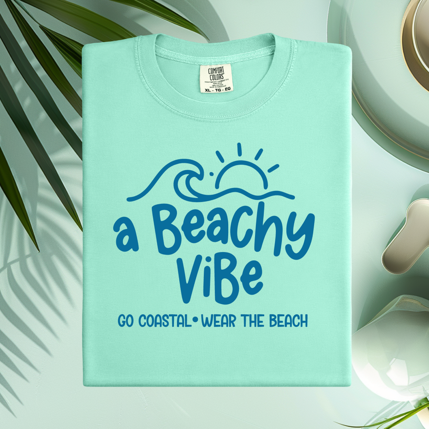 Light blue t-shirt with 'a Beachy Vibe' text on a white surface with green leaves.