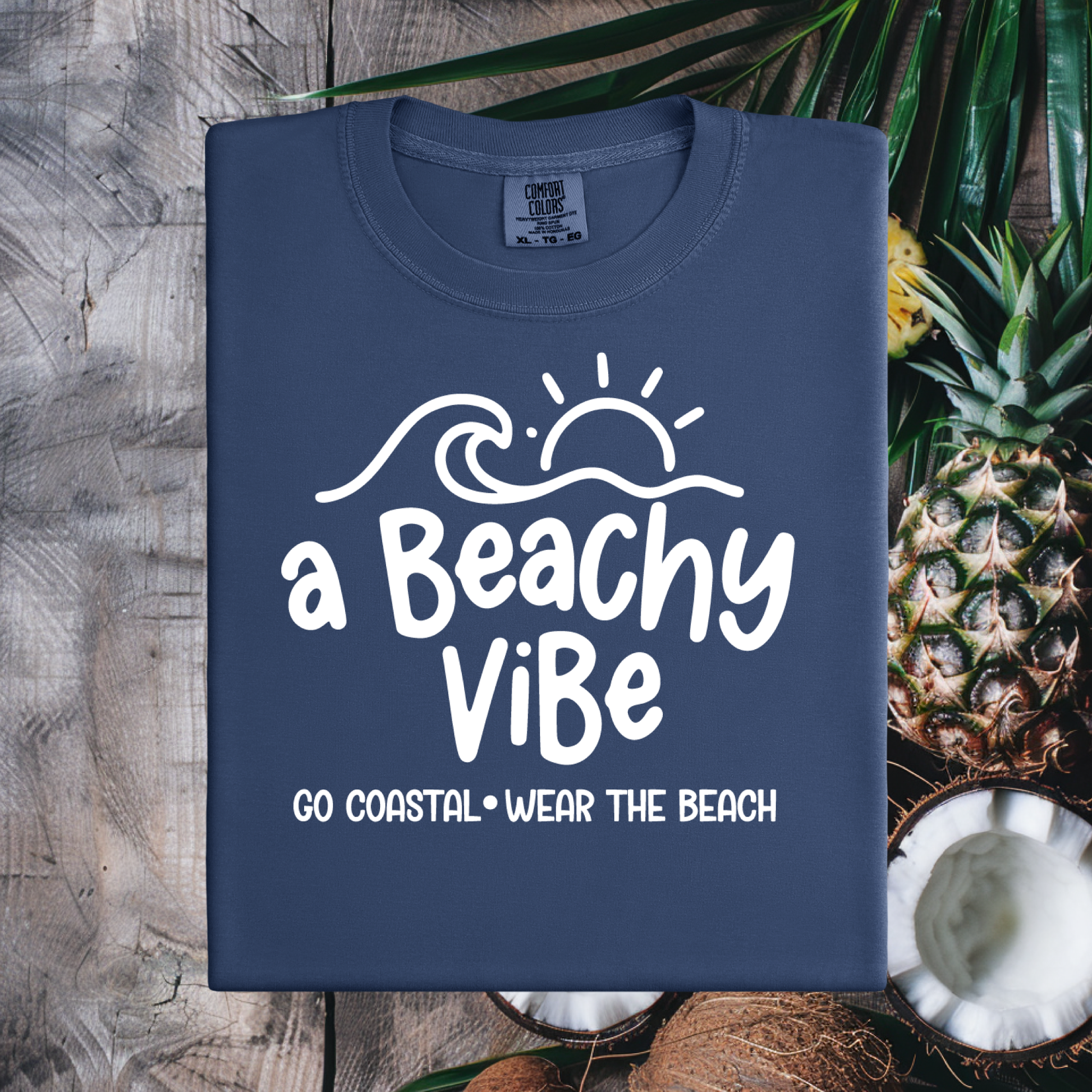 Navy blue t-shirt with 'a Beachy Vibe' text on a wooden surface with tropical elements.