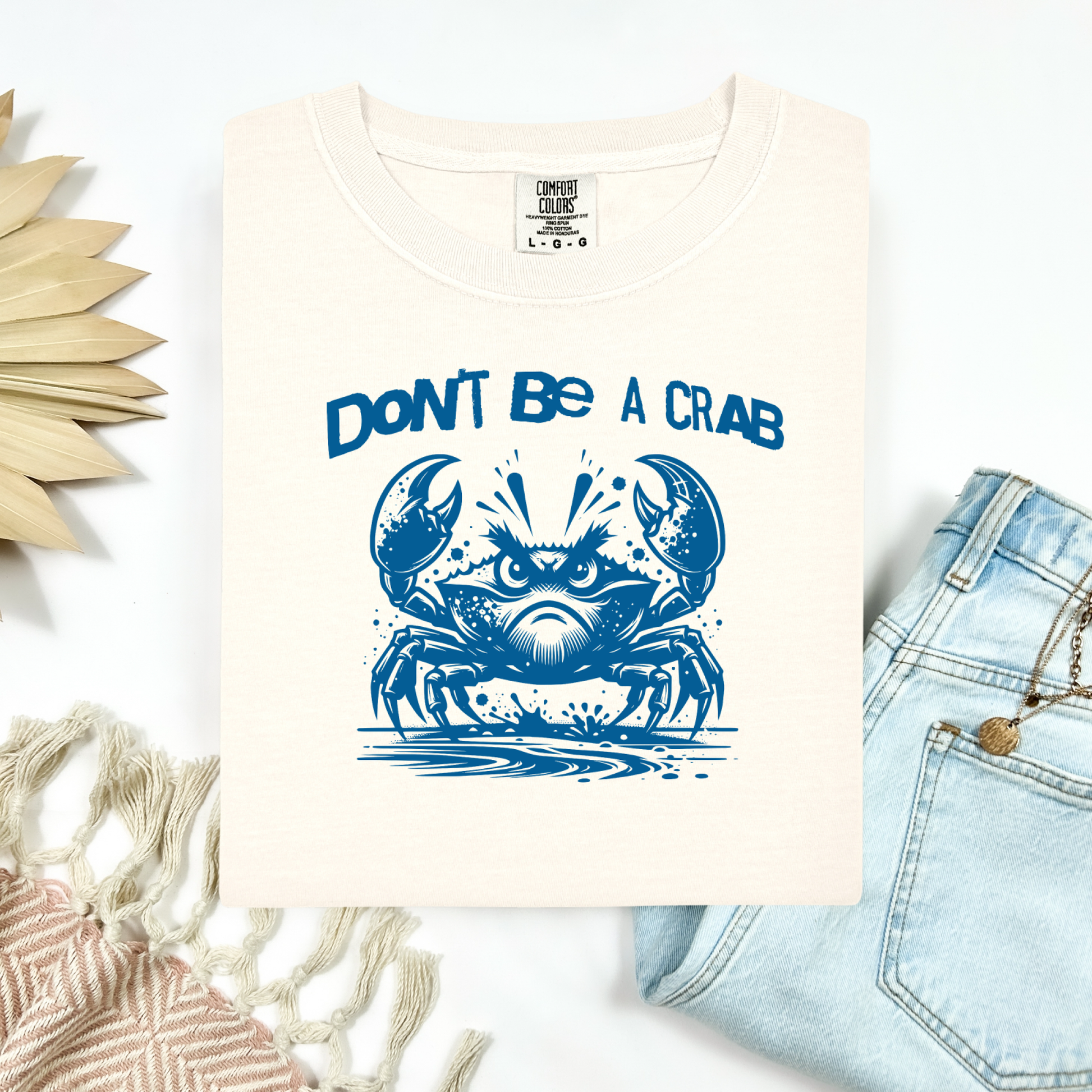 Comfort Colors 1717 "Don't Be A Crab" t shirt with cute crab looking angry with  in a  one color graphic. Picture on a flay lay mockup with pair of blue jeans and blanket and a wicker sun shaped accessory.
