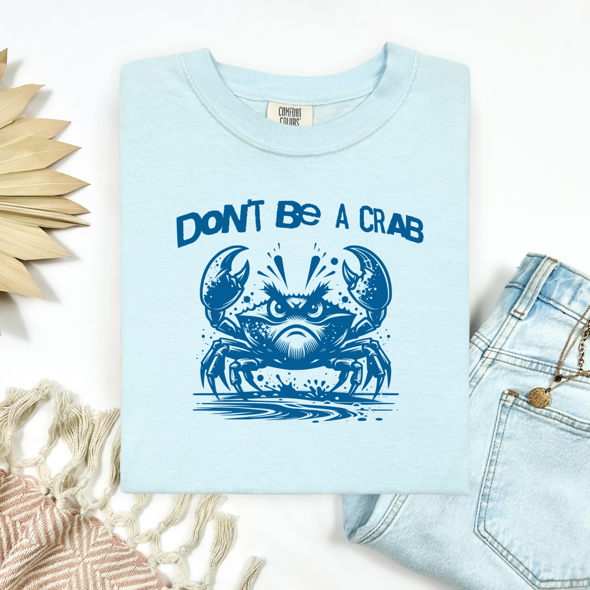Comfort Colors 1717 "Don't Be A Crab" t shirt with cute crab looking angry with  in a  one color graphic. Picture on a flay lay mockup with pair of blue jeans and blanket and a wicker sun shaped accessory.