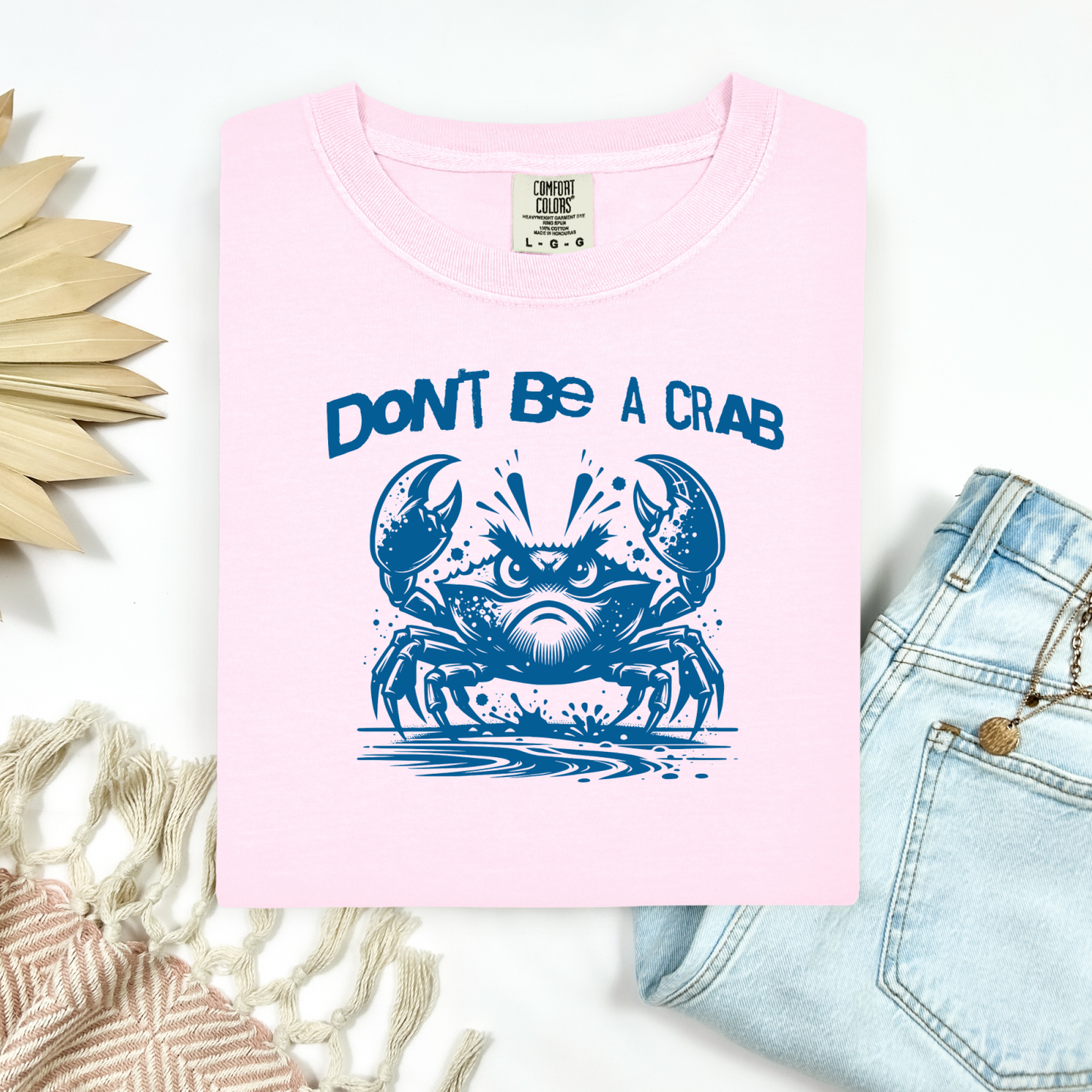 Comfort Colors 1717 "Don't Be A Crab" t shirt with cute crab looking angry with  in a  one color graphic. Picture on a flay lay mockup with pair of blue jeans and blanket and a wicker sun shaped accessory.