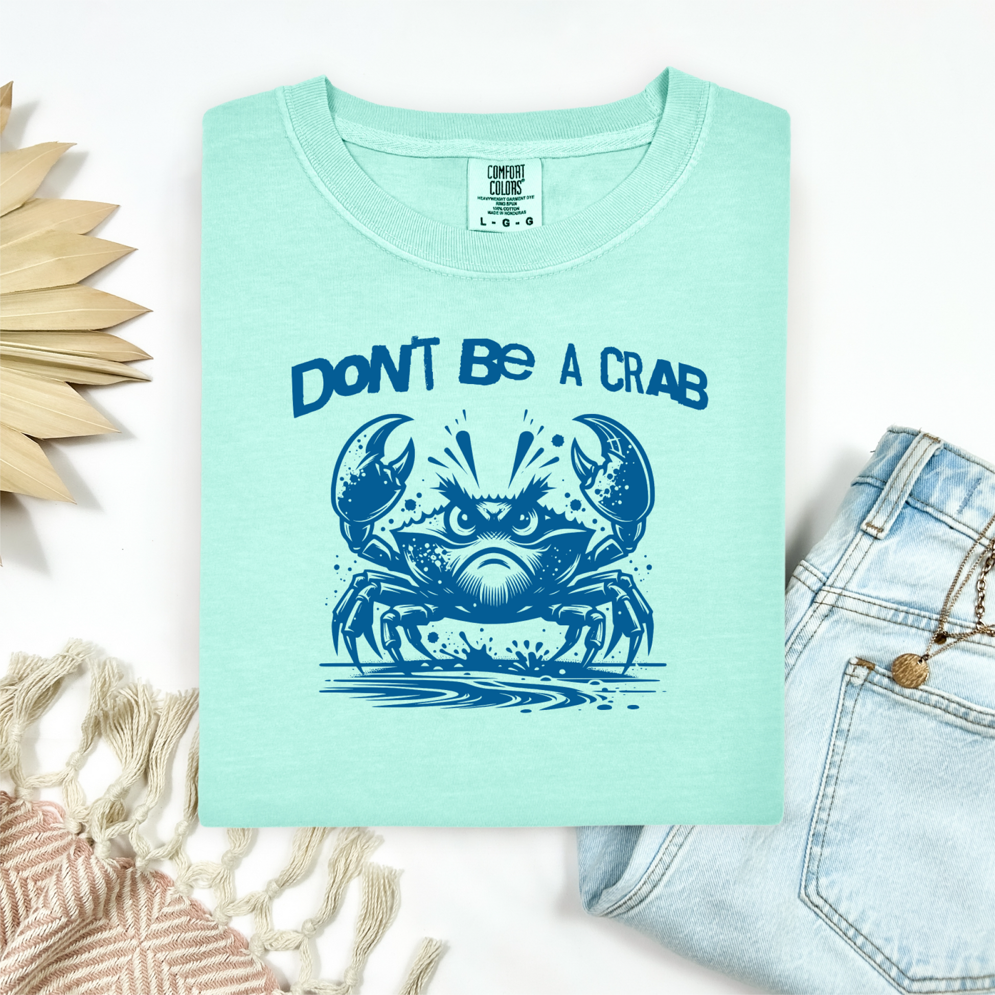 Comfort Colors 1717 "Don't Be A Crab" t shirt with cute crab looking angry with  in a  one color graphic. Picture on a flay lay mockup with pair of blue jeans and blanket and a wicker sun shaped accessory.