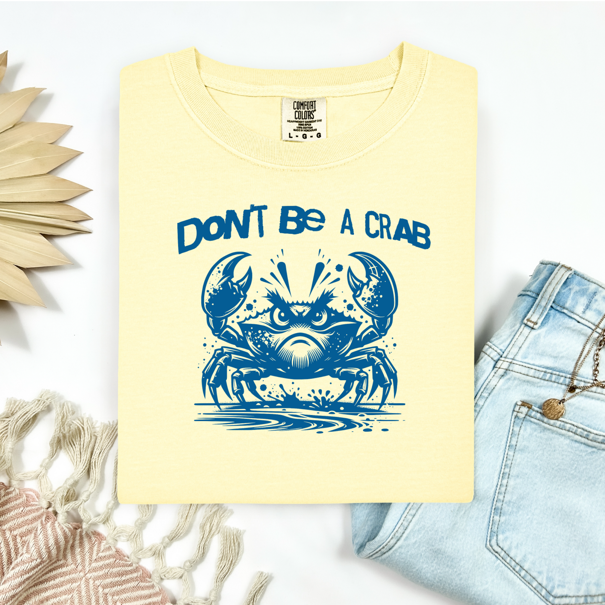 Comfort Colors 1717 "Don't Be A Crab" t shirt with cute crab looking angry with  in a  one color graphic. Picture on a flay lay mockup with pair of blue jeans and blanket and a wicker sun shaped accessory.