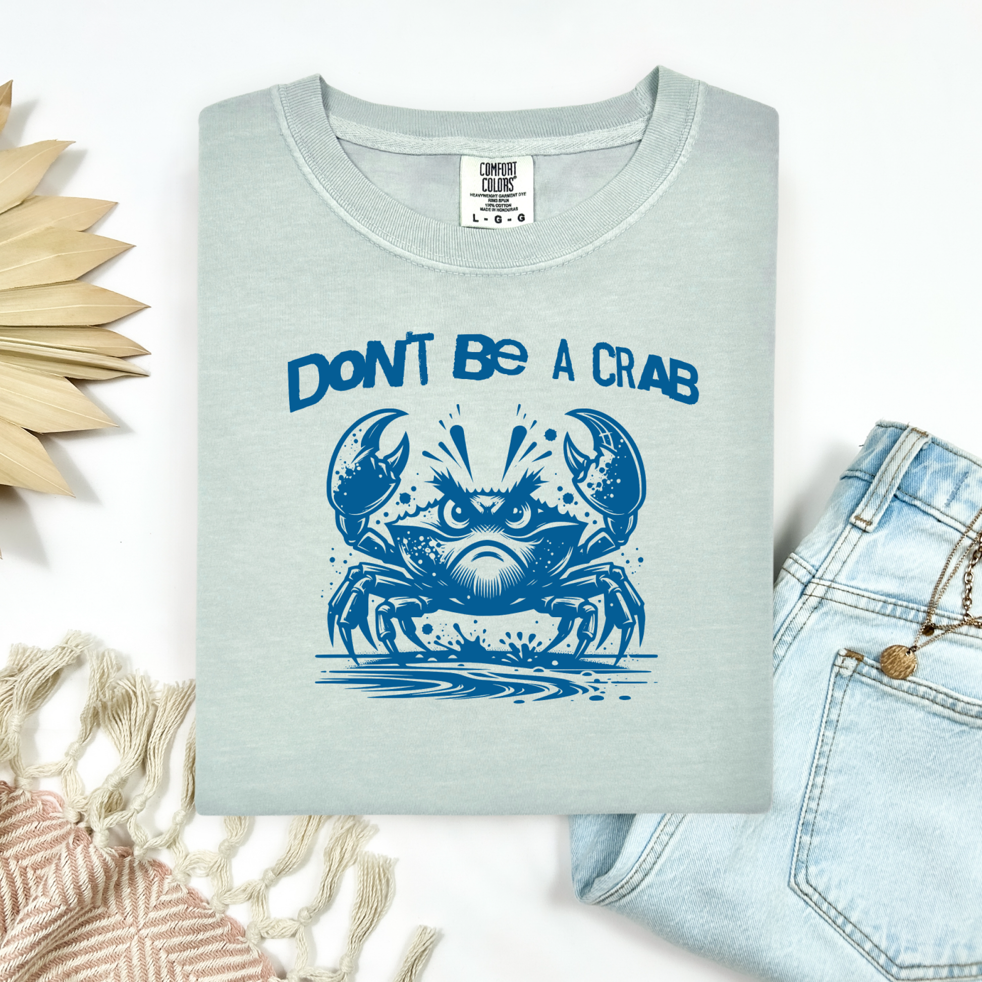Comfort Colors 1717 "Don't Be A Crab" t shirt with cute crab looking angry with  in a  one color graphic. Picture on a flay lay mockup with pair of blue jeans and blanket and a wicker sun shaped accessory.
