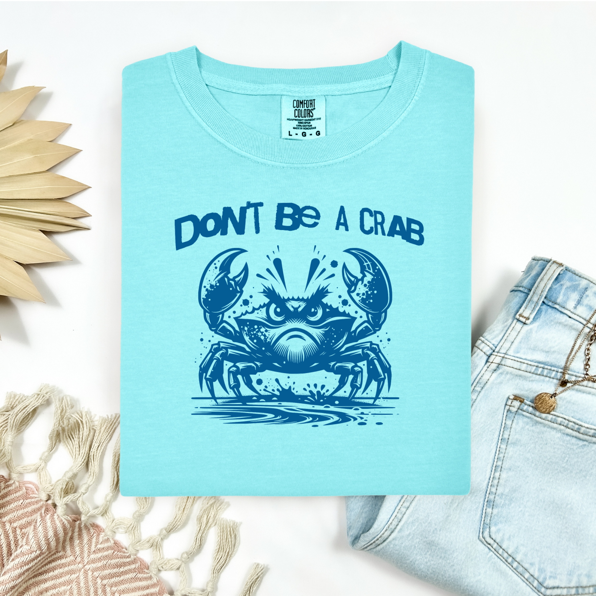 Comfort Colors 1717 "Don't Be A Crab" t shirt with cute crab looking angry with  in a  one color graphic. Picture on a flay lay mockup with pair of blue jeans and blanket and a wicker sun shaped accessory.
