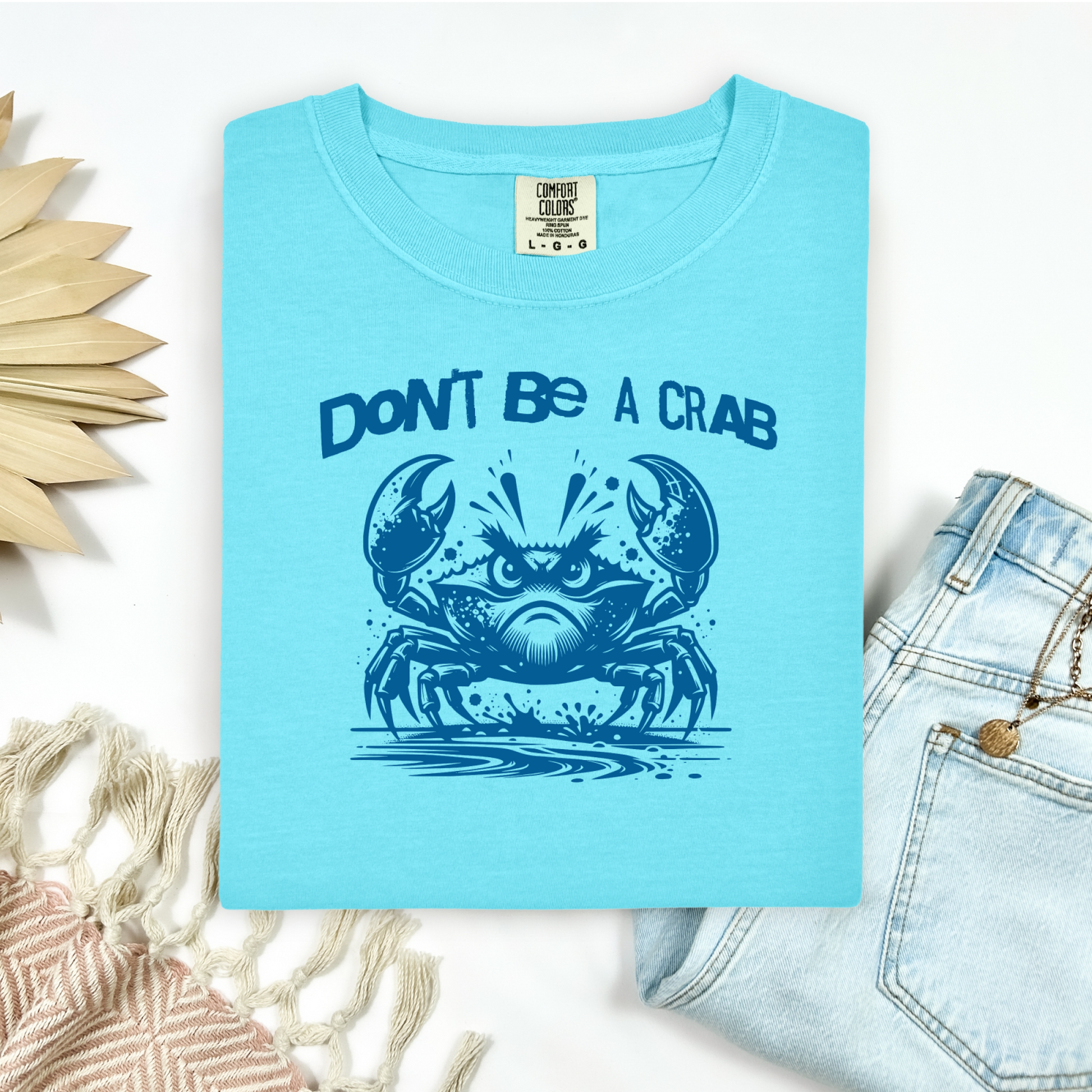 Comfort Colors 1717 "Don't Be A Crab" t shirt with cute crab looking angry with  in a  one color graphic. Picture on a flay lay mockup with pair of blue jeans and blanket and a wicker sun shaped accessory.