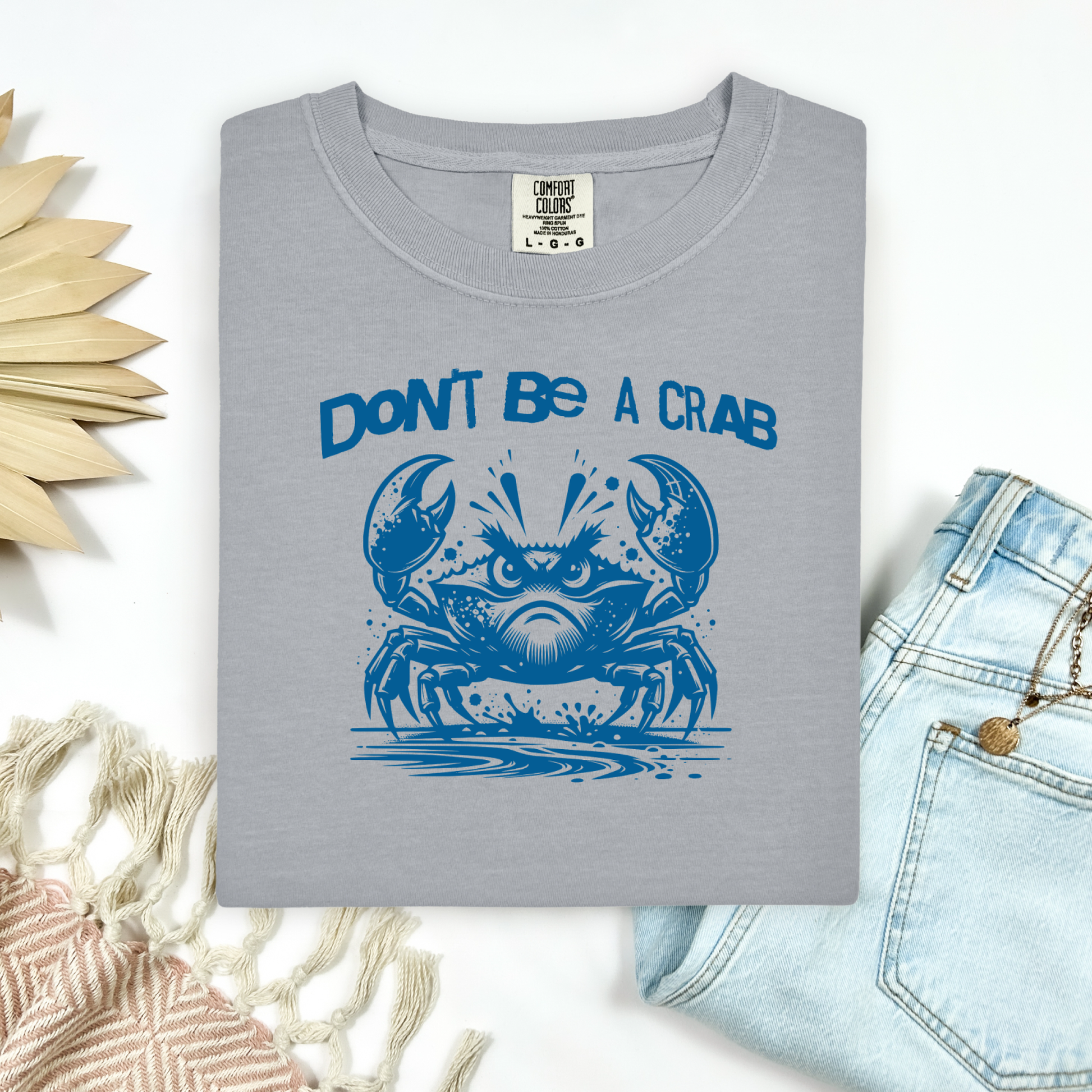 Comfort Colors 1717 "Don't Be A Crab" t shirt with cute crab looking angry with  in a  one color graphic. Picture on a flay lay mockup with pair of blue jeans and blanket and a wicker sun shaped accessory.