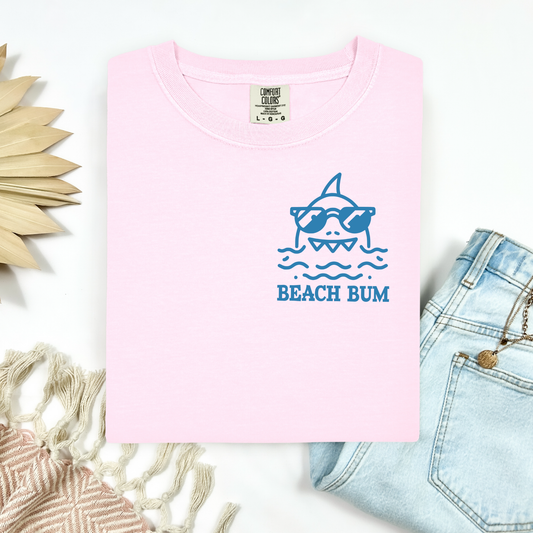 Comfort Colors 1717 "Beach Bum" t shirt with a simple line drawing of a shark wearing sunglasses in a one color graphic. Picture on a flay lay mockup with pair of blue jeans and blanket and a wicker sun shaped accessory.