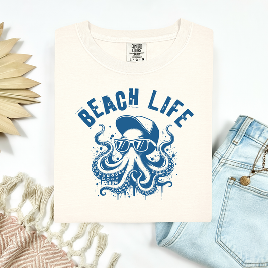 Comfort Colors 1717 "Beach Life" t shirt with a octopus with a baseball cap and sunglasses in a one color graphic. Picture on a flay lay mockup with pair of blue jeans and blanket and a wicker sun shaped accessory.
