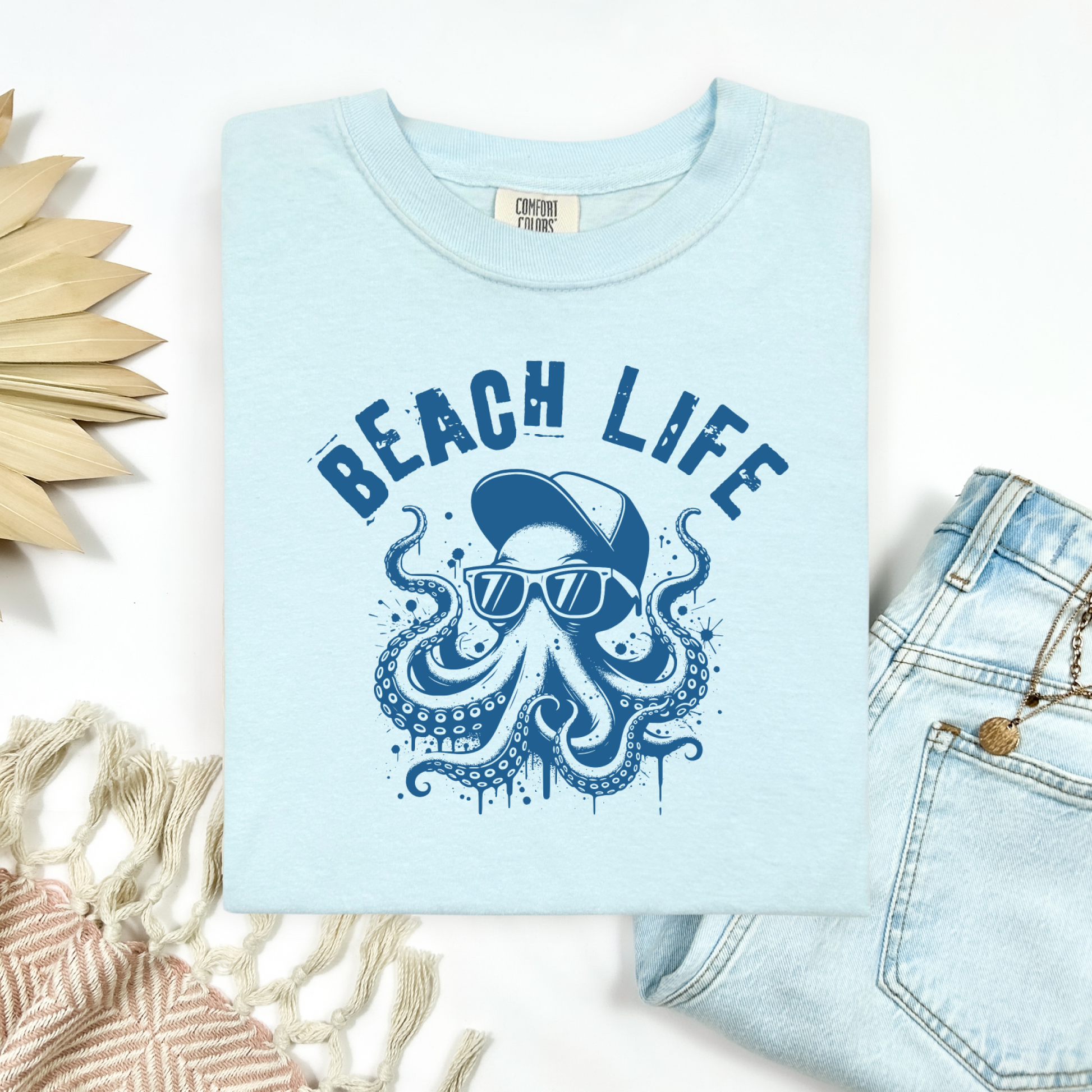 Comfort Colors 1717 "Beach Life" t shirt with a octopus with a baseball cap and sunglasses in a one color graphic. Picture on a flay lay mockup with pair of blue jeans and blanket and a wicker sun shaped accessory.