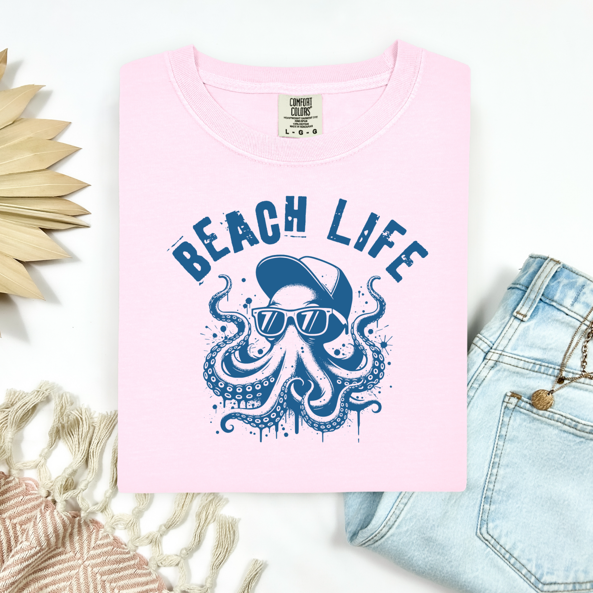 Comfort Colors 1717 "Beach Life" t shirt with a octopus with a baseball cap and sunglasses in a one color graphic. Picture on a flay lay mockup with pair of blue jeans and blanket and a wicker sun shaped accessory.
