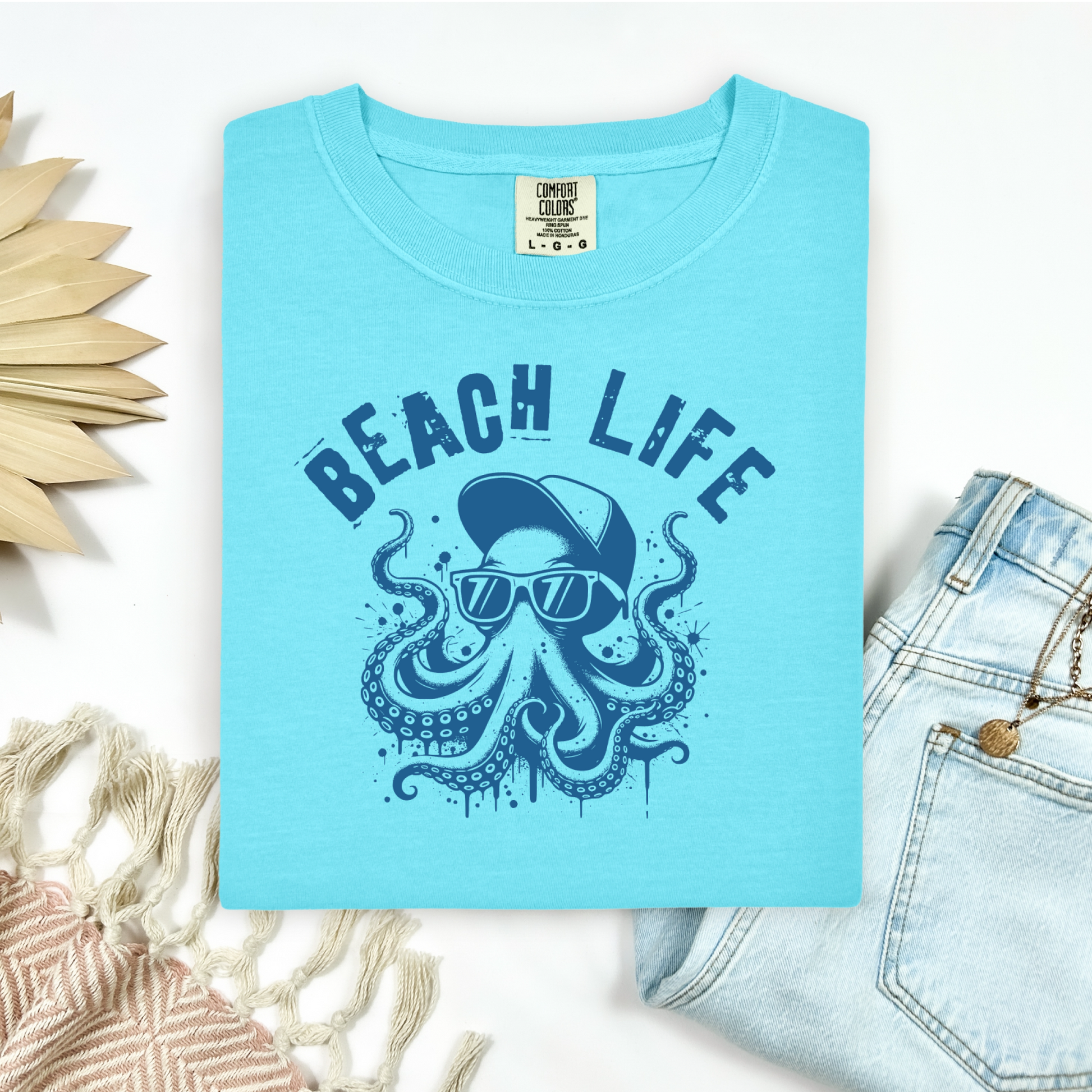 Comfort Colors 1717 "Beach Life" t shirt with a octopus with a baseball cap and sunglasses in a one color graphic. Picture on a flay lay mockup with pair of blue jeans and blanket and a wicker sun shaped accessory.