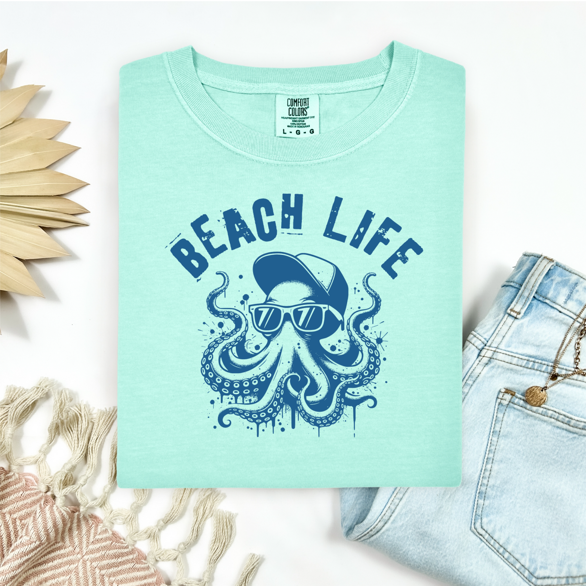 Comfort Colors 1717 "Beach Life" t shirt with a octopus with a baseball cap and sunglasses in a one color graphic. Picture on a flay lay mockup with pair of blue jeans and blanket and a wicker sun shaped accessory.