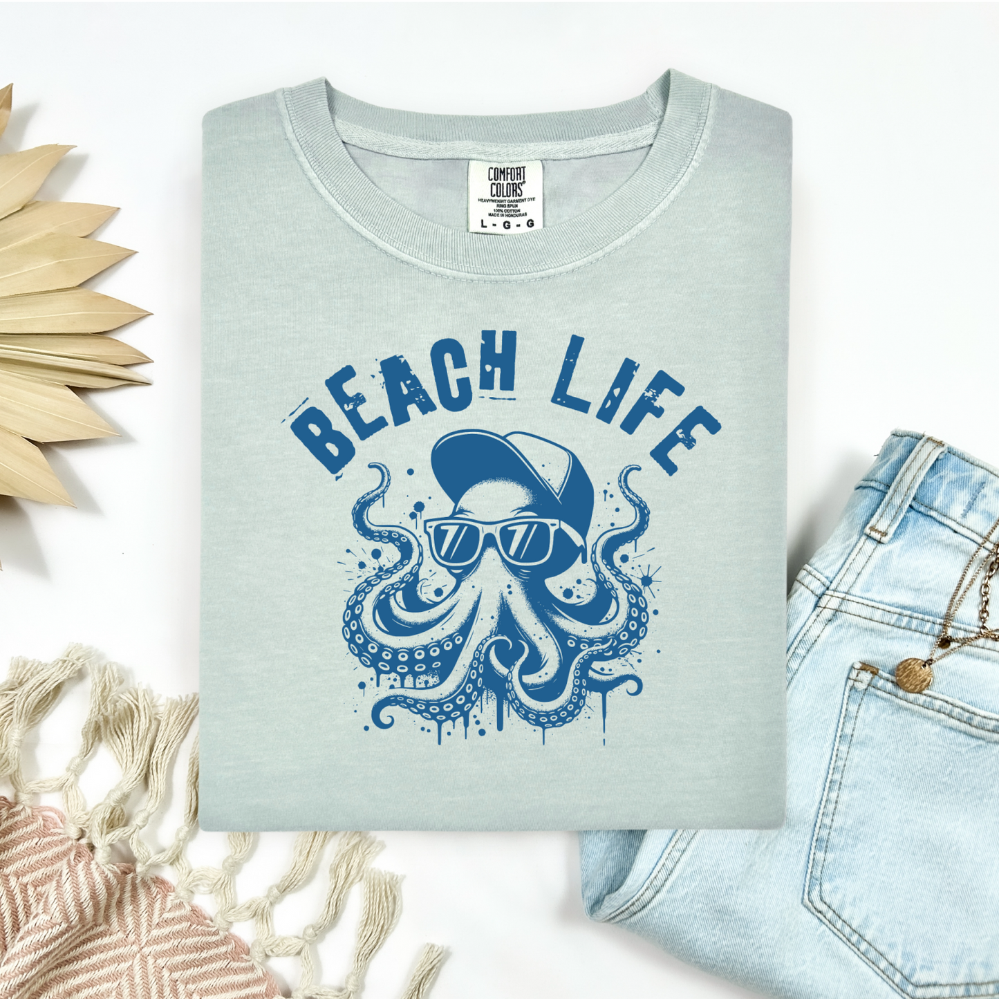 Comfort Colors 1717 "Beach Life" t shirt with a octopus with a baseball cap and sunglasses in a one color graphic. Picture on a flay lay mockup with pair of blue jeans and blanket and a wicker sun shaped accessory.