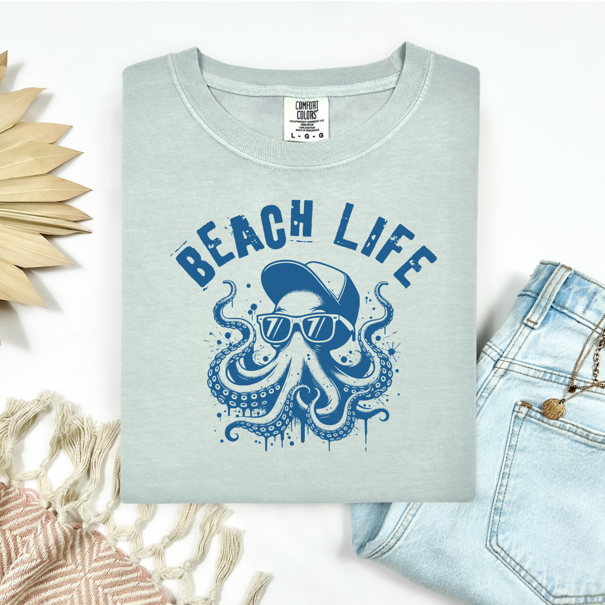 Comfort Colors 1717 "Beach Life" t shirt with a octopus with a baseball cap and sunglasses in a one color graphic. Picture on a flay lay mockup with pair of blue jeans and blanket and a wicker sun shaped accessory.