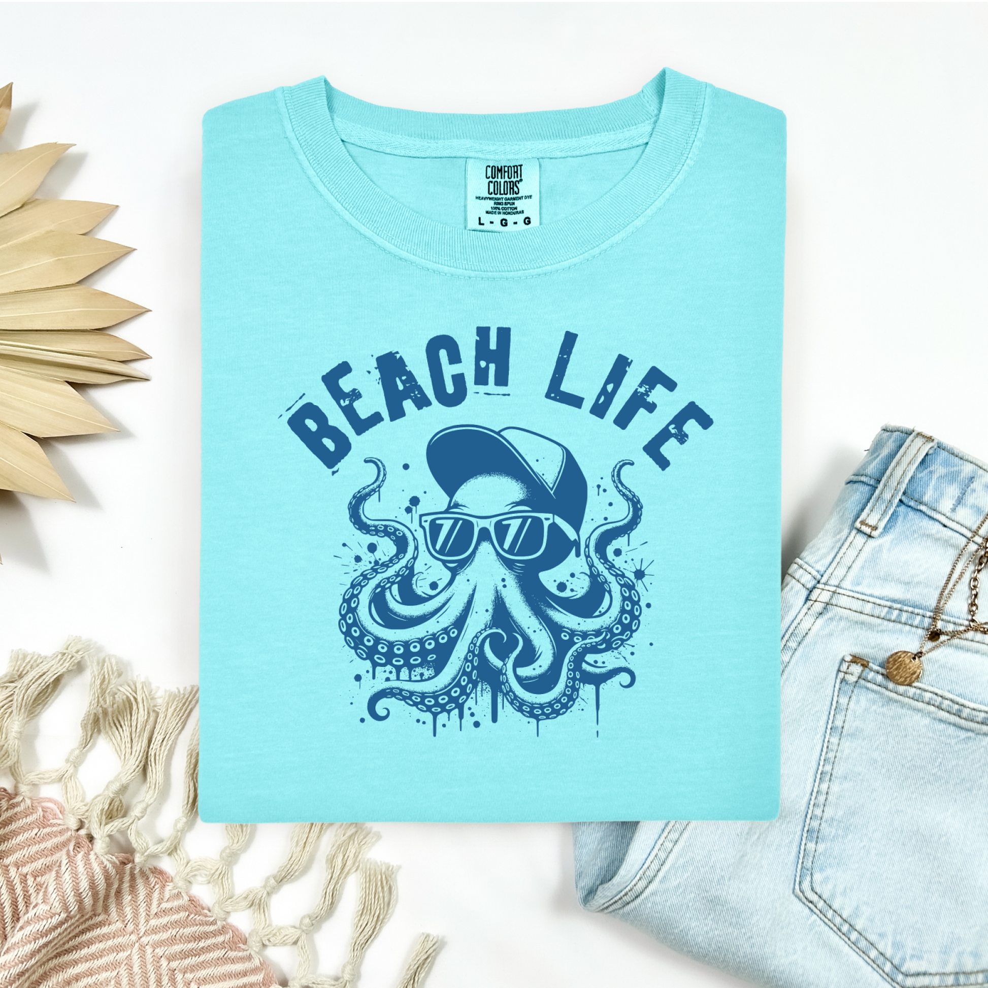 Comfort Colors 1717 "Beach Life" t shirt with a octopus with a baseball cap and sunglasses in a one color graphic. Picture on a flay lay mockup with pair of blue jeans and blanket and a wicker sun shaped accessory.