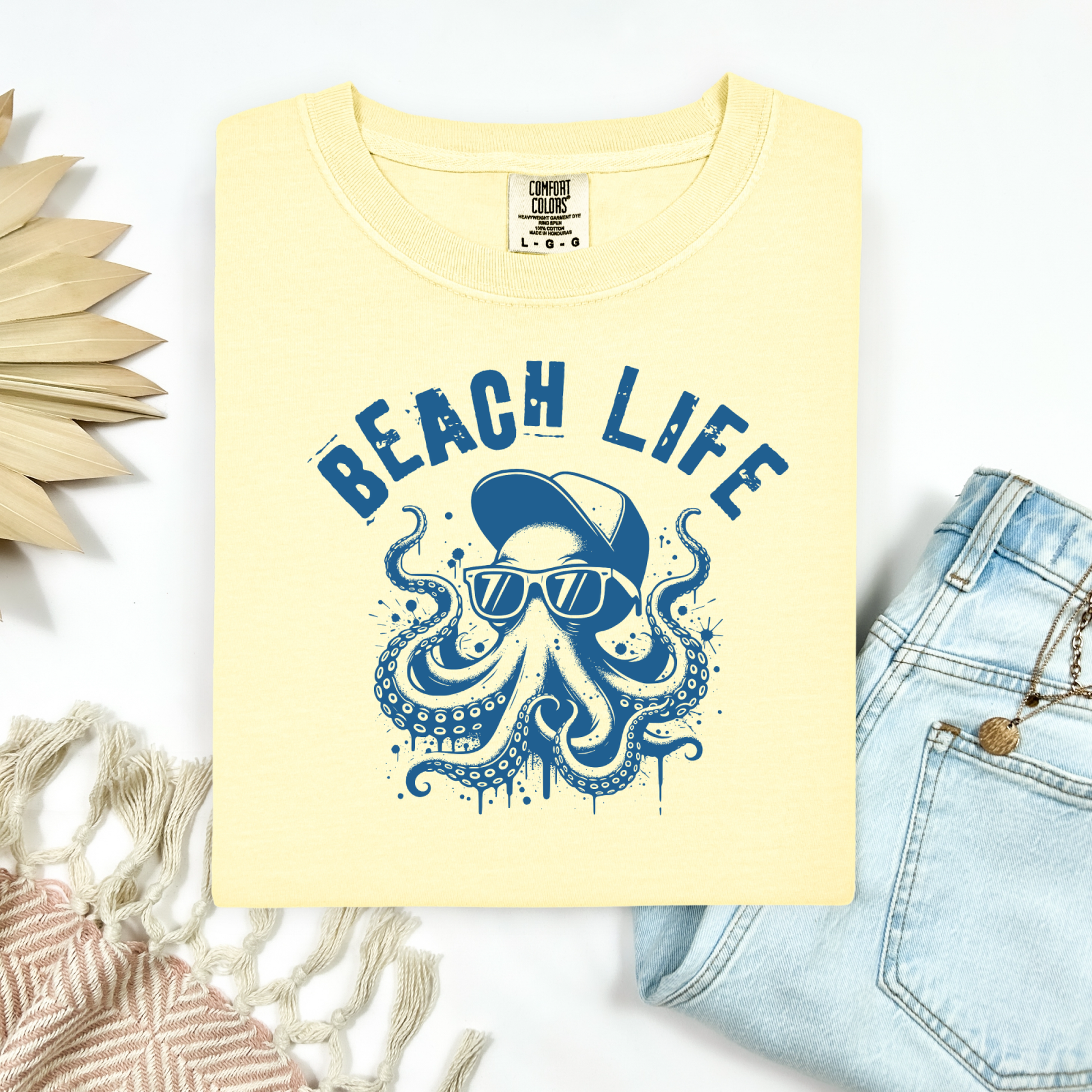 Comfort Colors 1717 "Beach Life" t shirt with a octopus with a baseball cap and sunglasses in a one color graphic. Picture on a flay lay mockup with pair of blue jeans and blanket and a wicker sun shaped accessory.