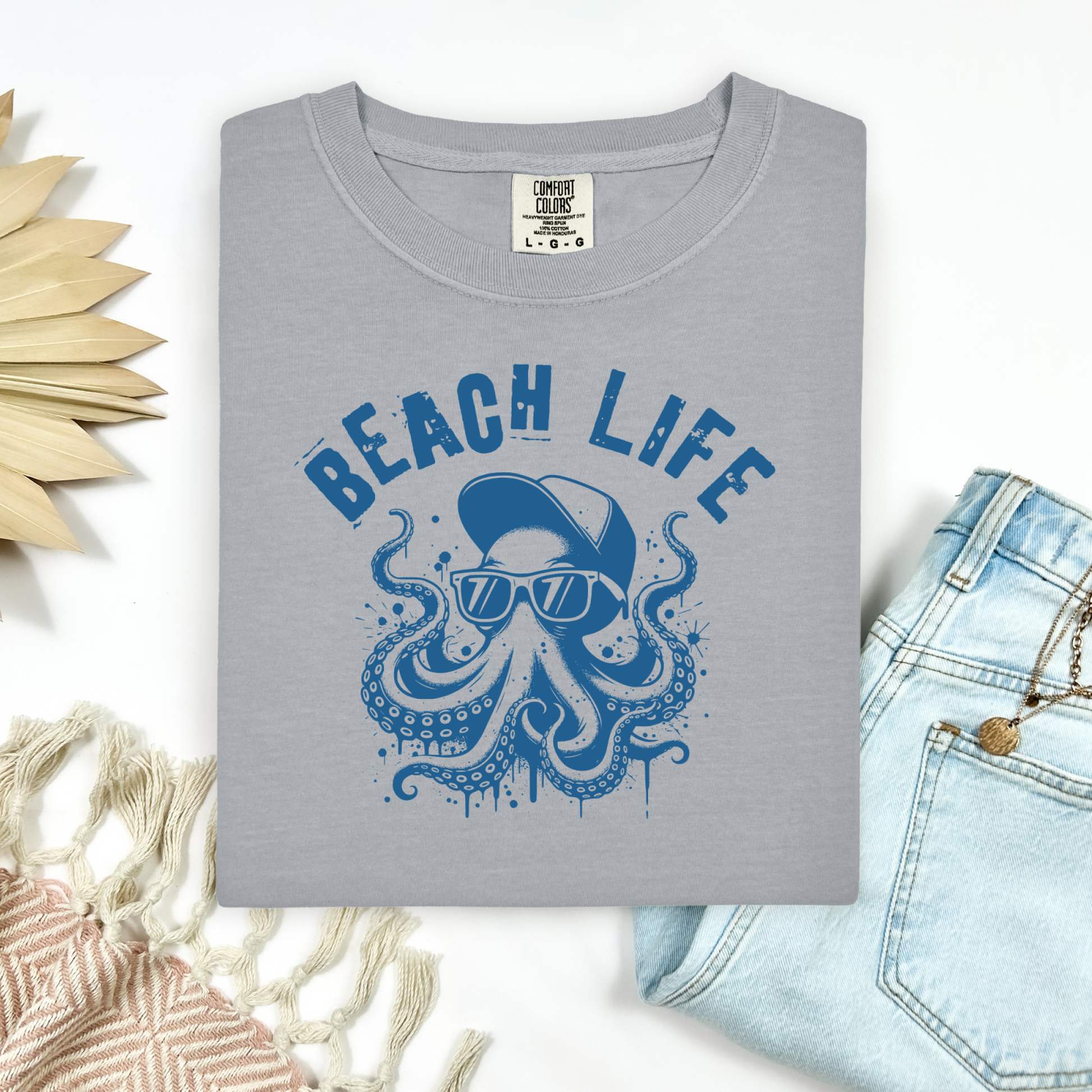 Comfort Colors 1717 "Beach Life" t shirt with a octopus with a baseball cap and sunglasses in a one color graphic. Picture on a flay lay mockup with pair of blue jeans and blanket and a wicker sun shaped accessory.
