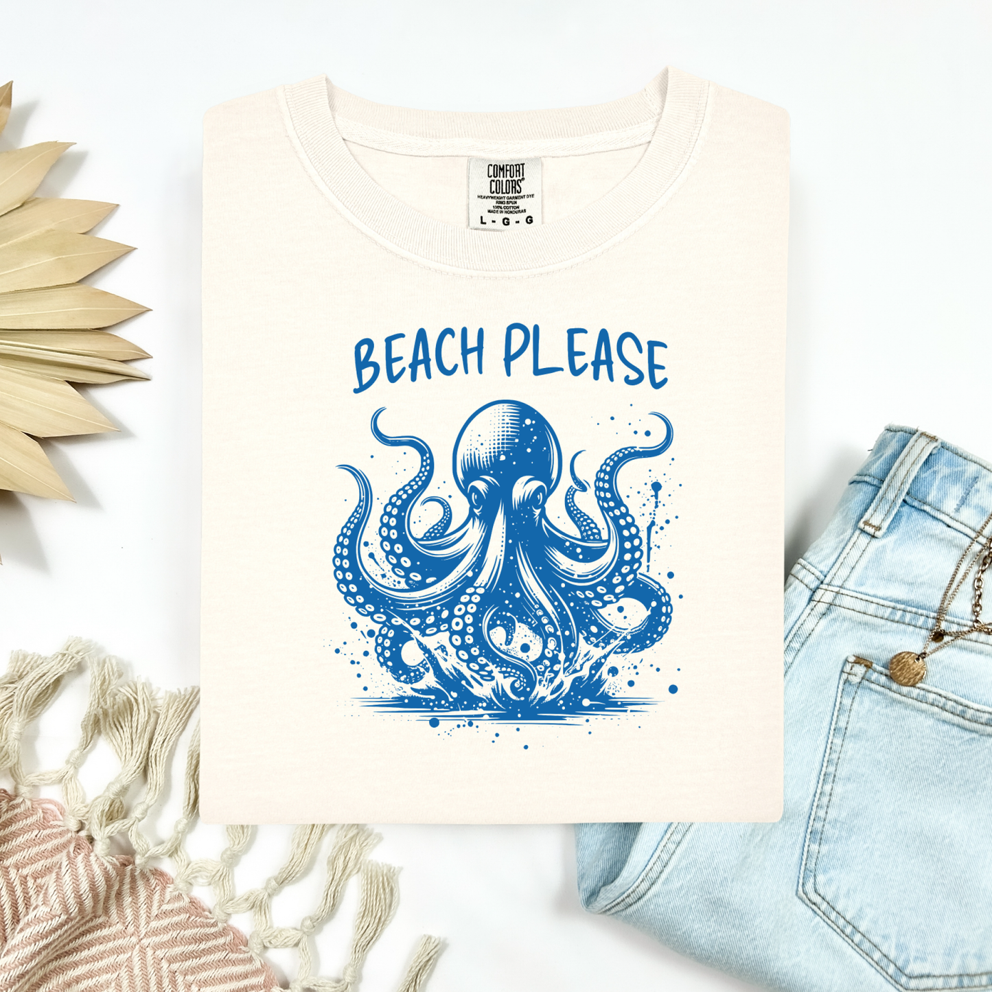 Comfort Colors 1717 "Beach Please" t shirt with a octopus  jumping in water in one color graphic. Picture on a flay lay mockup with pair of blue jeans and blanket and a wicker sun shaped accessory.