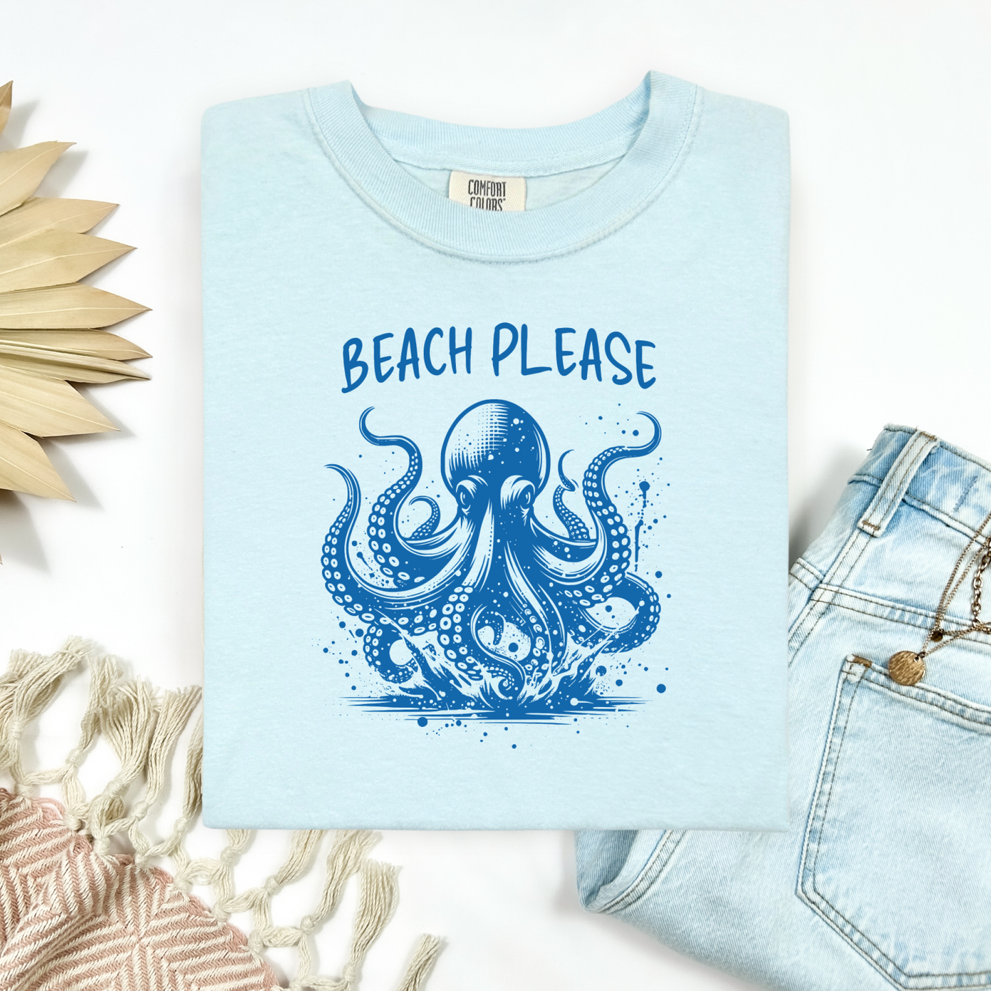 Comfort Colors 1717 "Beach Please" t shirt with a octopus  jumping in water in one color graphic. Picture on a flay lay mockup with pair of blue jeans and blanket and a wicker sun shaped accessory.