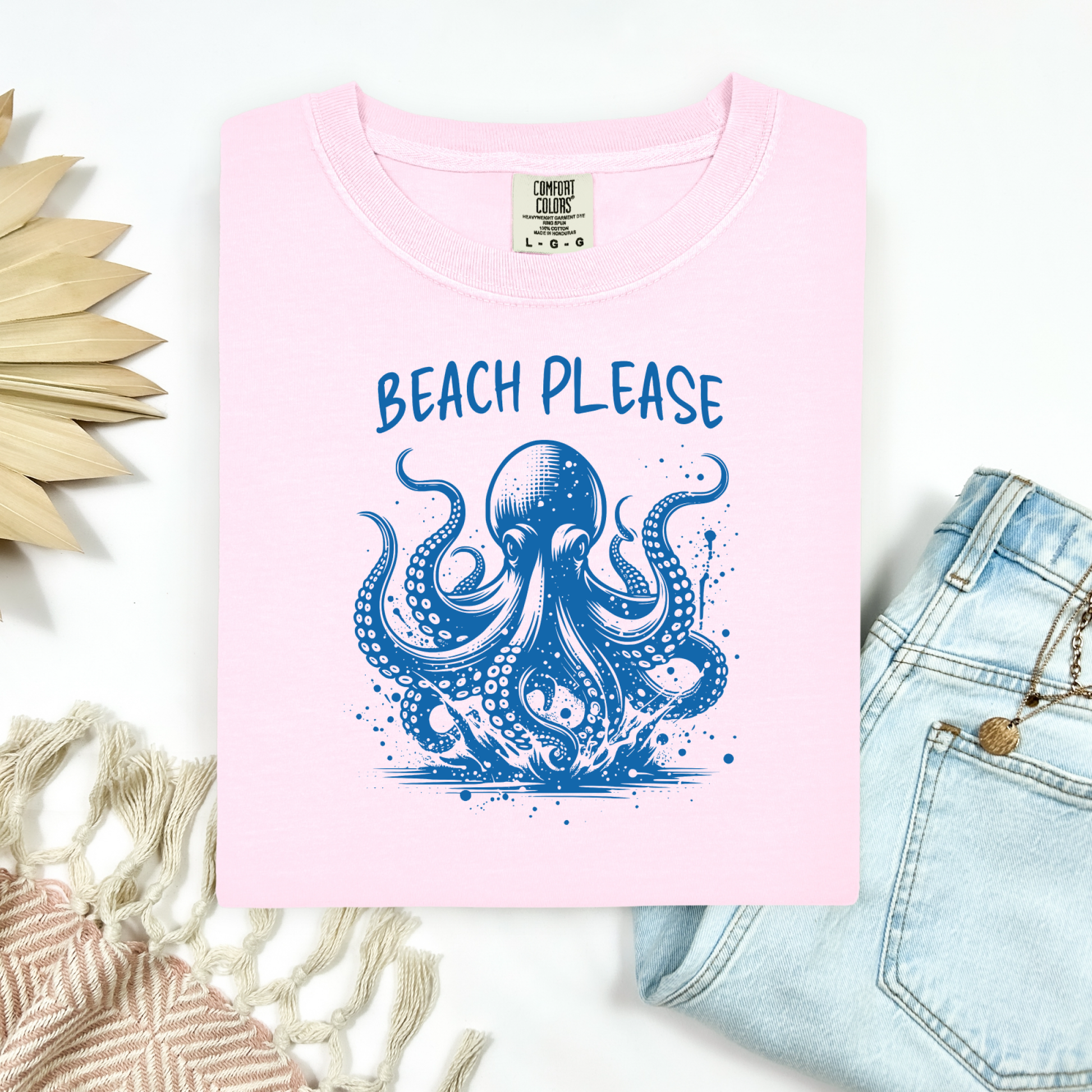 Comfort Colors 1717 "Beach Please" t shirt with a octopus  jumping in water in one color graphic. Picture on a flay lay mockup with pair of blue jeans and blanket and a wicker sun shaped accessory.