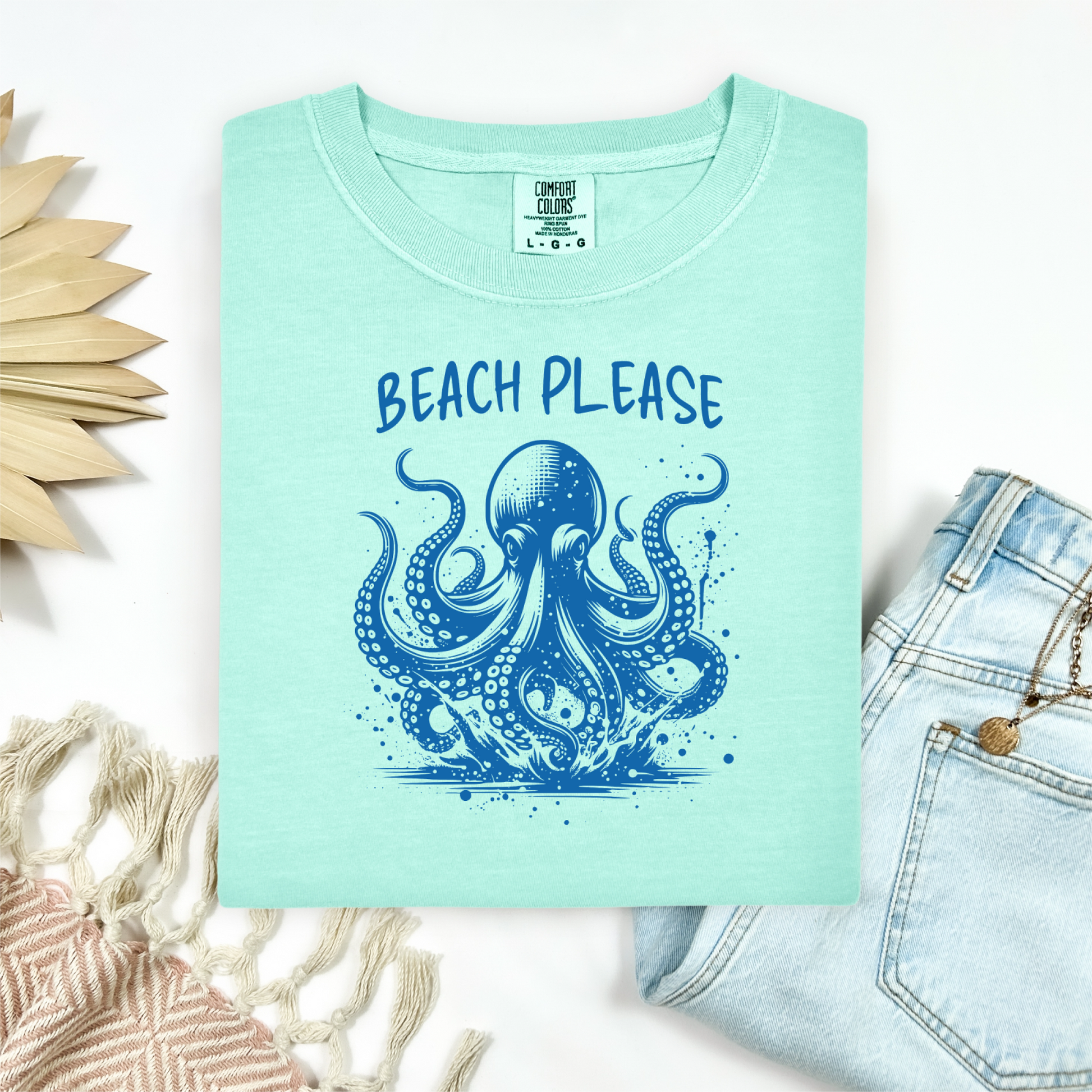 Comfort Colors 1717 "Beach Please" t shirt with a octopus  jumping in water in one color graphic. Picture on a flay lay mockup with pair of blue jeans and blanket and a wicker sun shaped accessory.