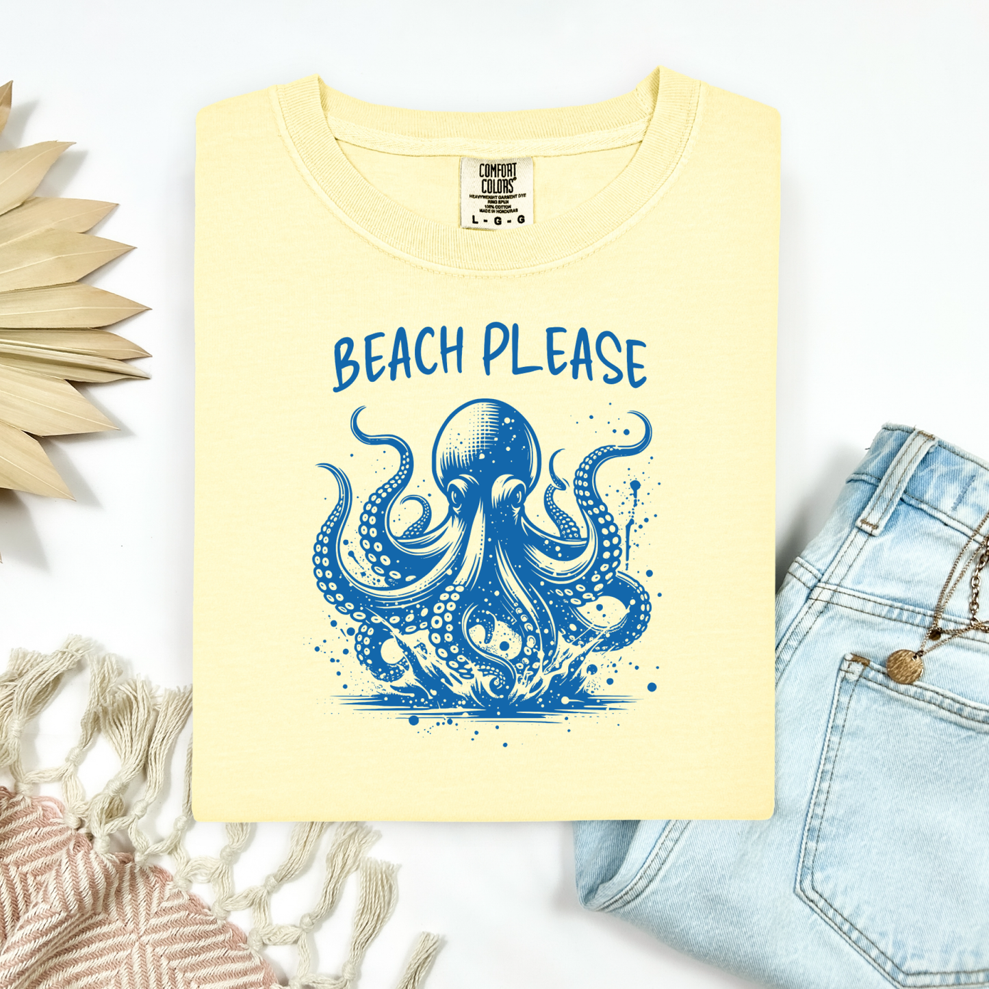 Comfort Colors 1717 "Beach Please" t shirt with a octopus  jumping in water in one color graphic. Picture on a flay lay mockup with pair of blue jeans and blanket and a wicker sun shaped accessory.