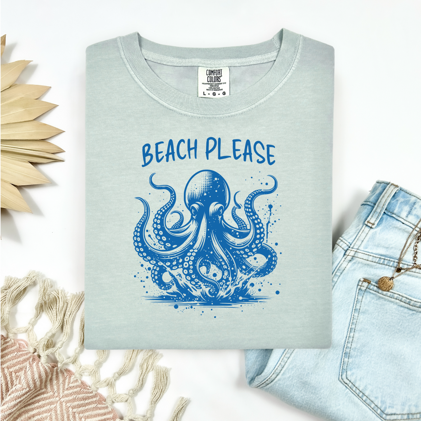 Comfort Colors 1717 "Beach Please" t shirt with a octopus  jumping in water in one color graphic. Picture on a flay lay mockup with pair of blue jeans and blanket and a wicker sun shaped accessory.