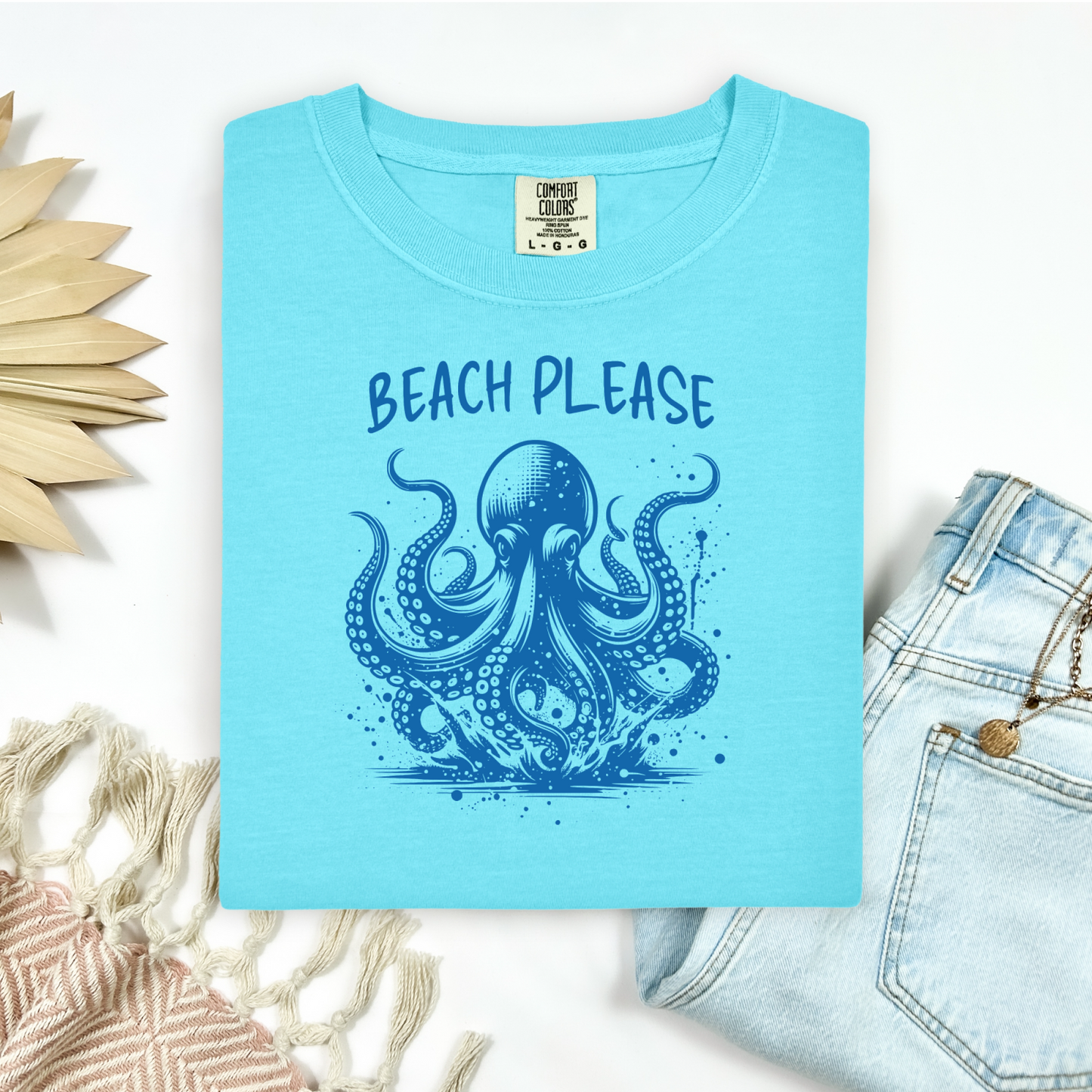 Comfort Colors 1717 "Beach Please" t shirt with a octopus  jumping in water in one color graphic. Picture on a flay lay mockup with pair of blue jeans and blanket and a wicker sun shaped accessory.
