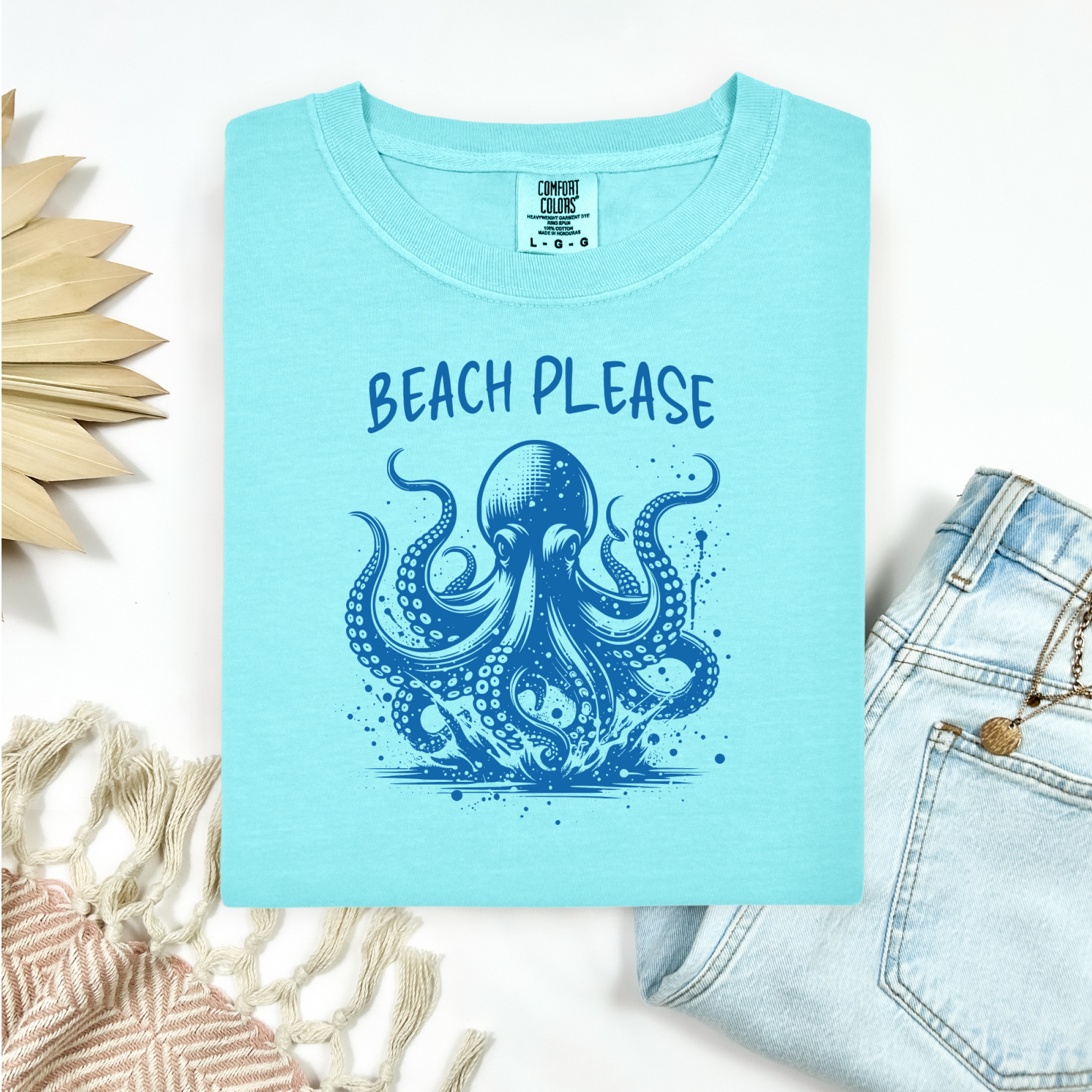 Comfort Colors 1717 "Beach Please" t shirt with a octopus  jumping in water in one color graphic. Picture on a flay lay mockup with pair of blue jeans and blanket and a wicker sun shaped accessory.