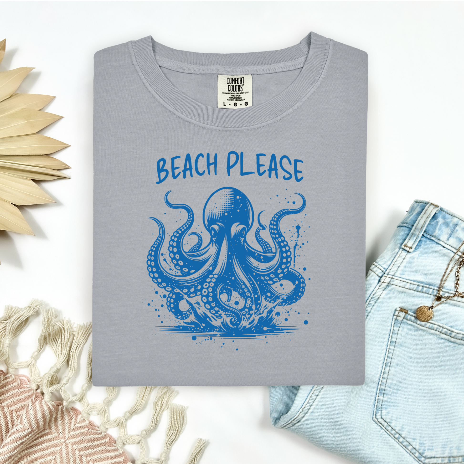 Comfort Colors 1717 "Beach Please" t shirt with a octopus  jumping in water in one color graphic. Picture on a flay lay mockup with pair of blue jeans and blanket and a wicker sun shaped accessory.